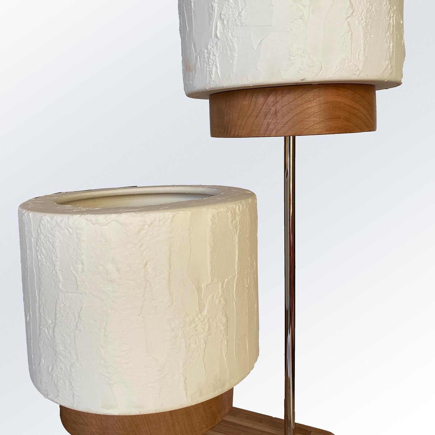 This sophisticated two-light table lamp couples a distinct sculptural flair with a striking illumination potential. Two thin metal rods in different heights connect the base in solid cherry marked by a ridged texture to the shading units, which are