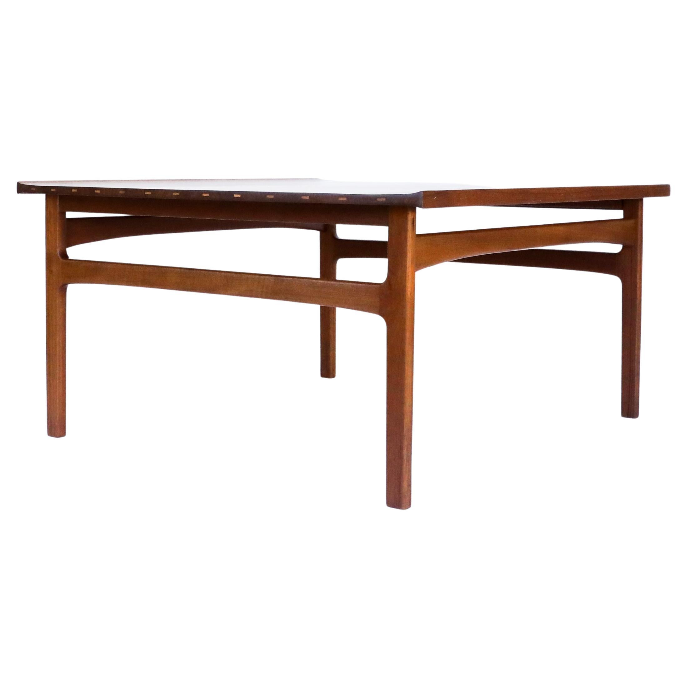 This is a stunning solid teak Danish coffee table which features a sculptural lipped edge top with two elegantly curved sides. The exposed wood joinery on the curved edges features distinctive inlays in a lighter, contrasting beech wood. This is an