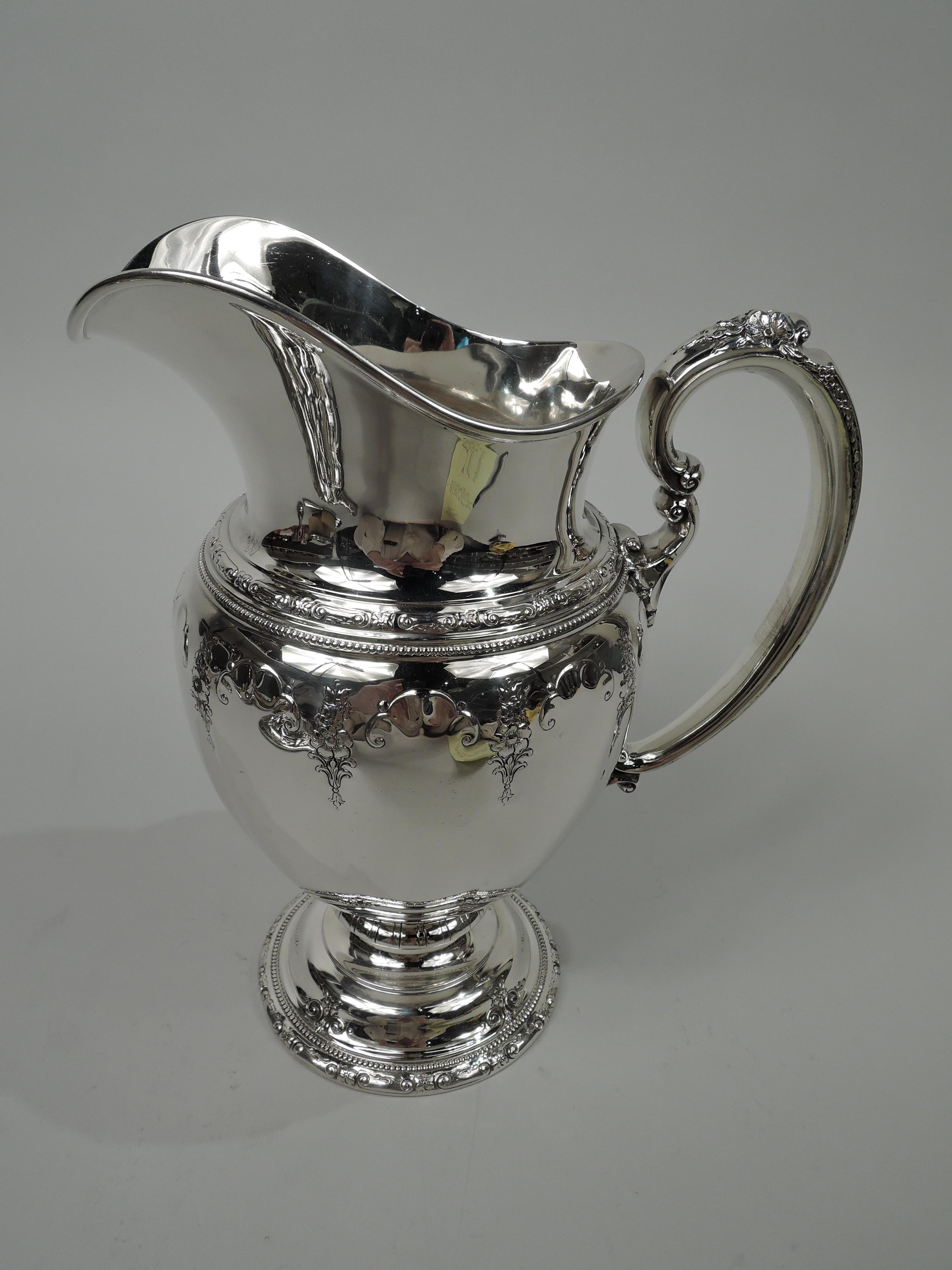Royal Windsor sterling silver water pitcher. Made by Towle in Newburyport, Mass. Ovoid bowl with helmet mouth, flower-capped double scroll handle, and domed foot. Chased and scalloped frames (vacant) interspersed with pendant flowers. Shoulder and