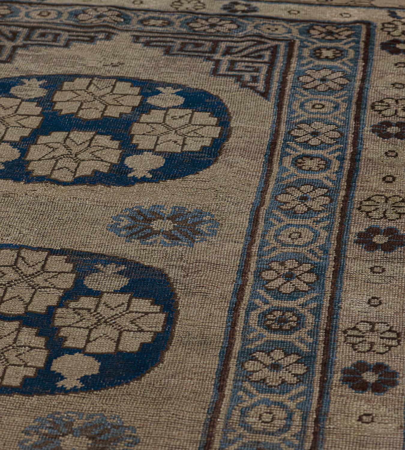 This traditional Khotan runner features a buff-brown field with a column of three royal-blue roundels, each containing buff-brown lozenges and stylized fruits, the roundels separated by paired light blue hooked rosettes, the mole-brown stepped