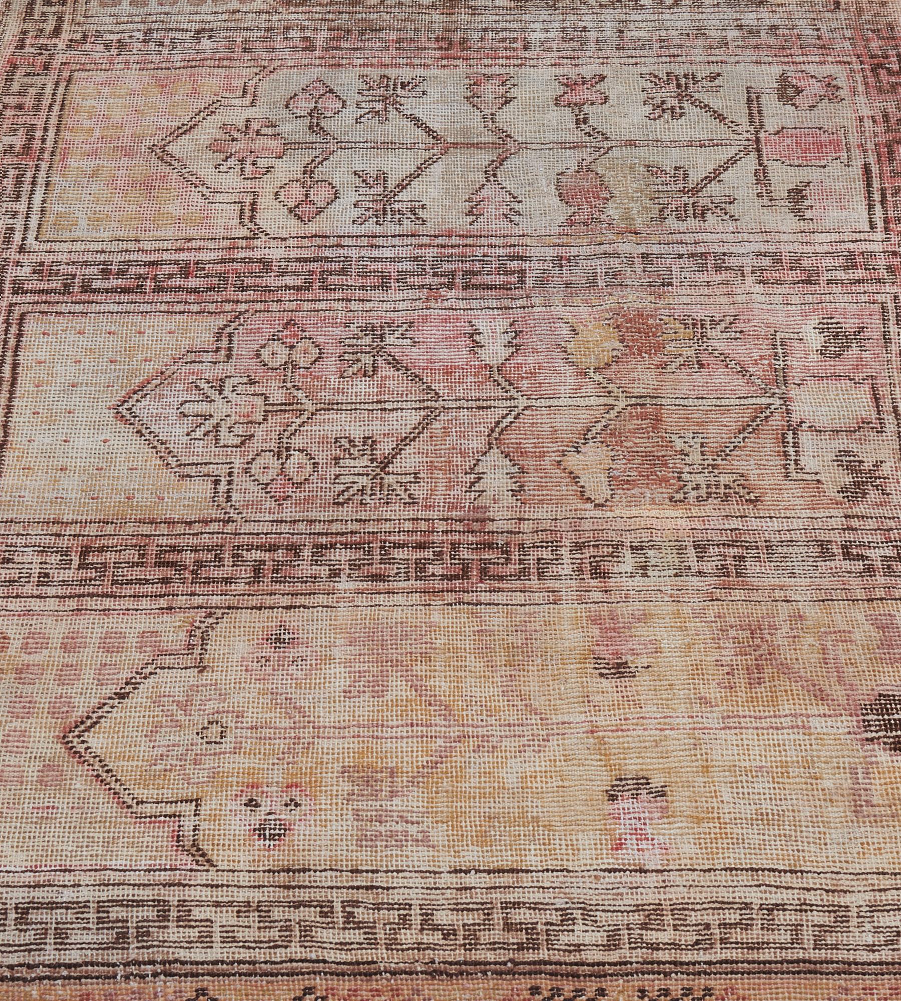 Ce tapis antique du Khotan, datant d'environ 1900, présente un champ rouge nuancé avec un motif de clé brun chocolat autour de six panneaux rectangulaires polychromes contenant chacun une grande tige fleurie angulaire, le panneau arqué au-dessus