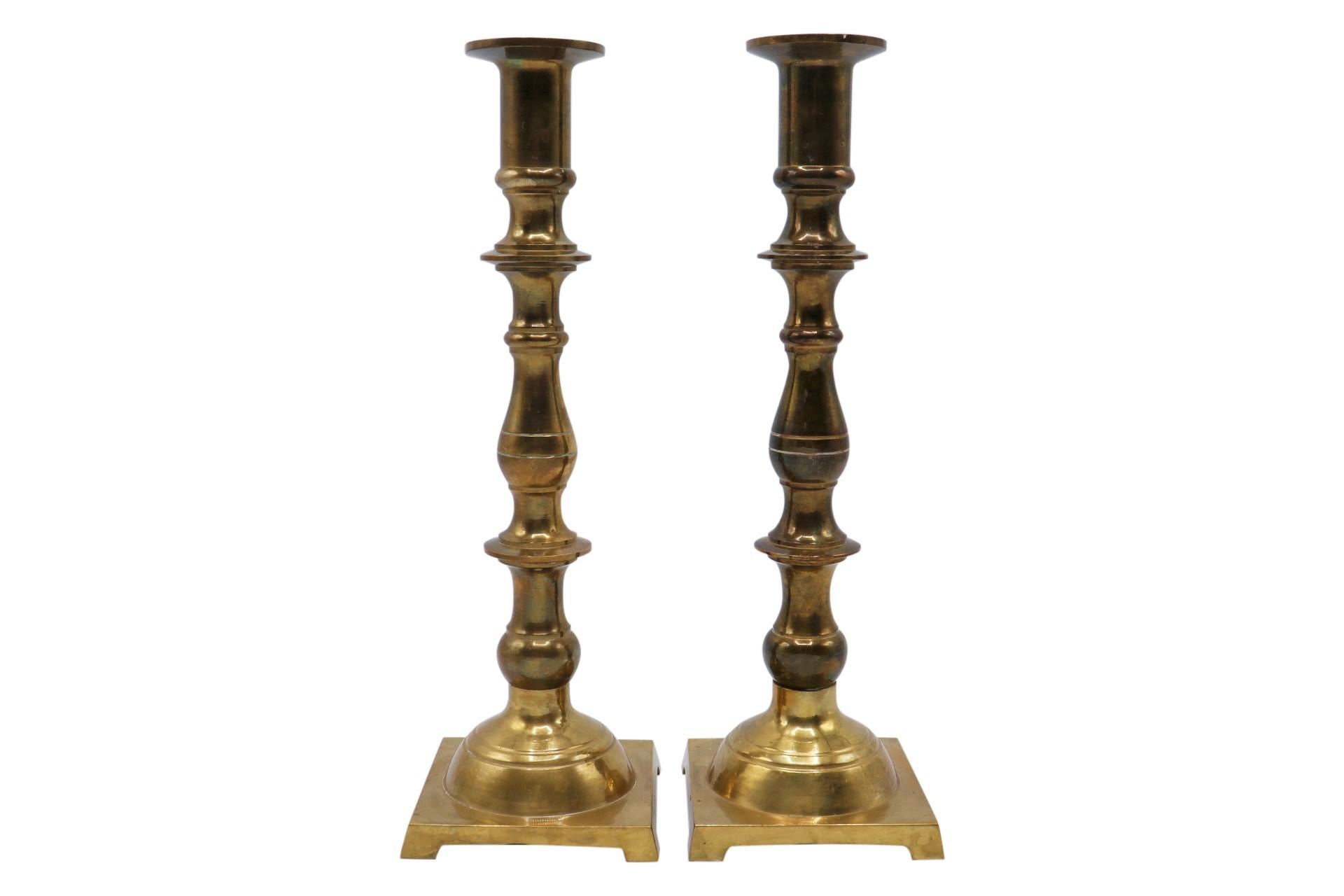 A pair of traditional turned candlesticks made of brass. Simple capitals top turned stems finished with domes above square feet. Dimensions per candlestick.