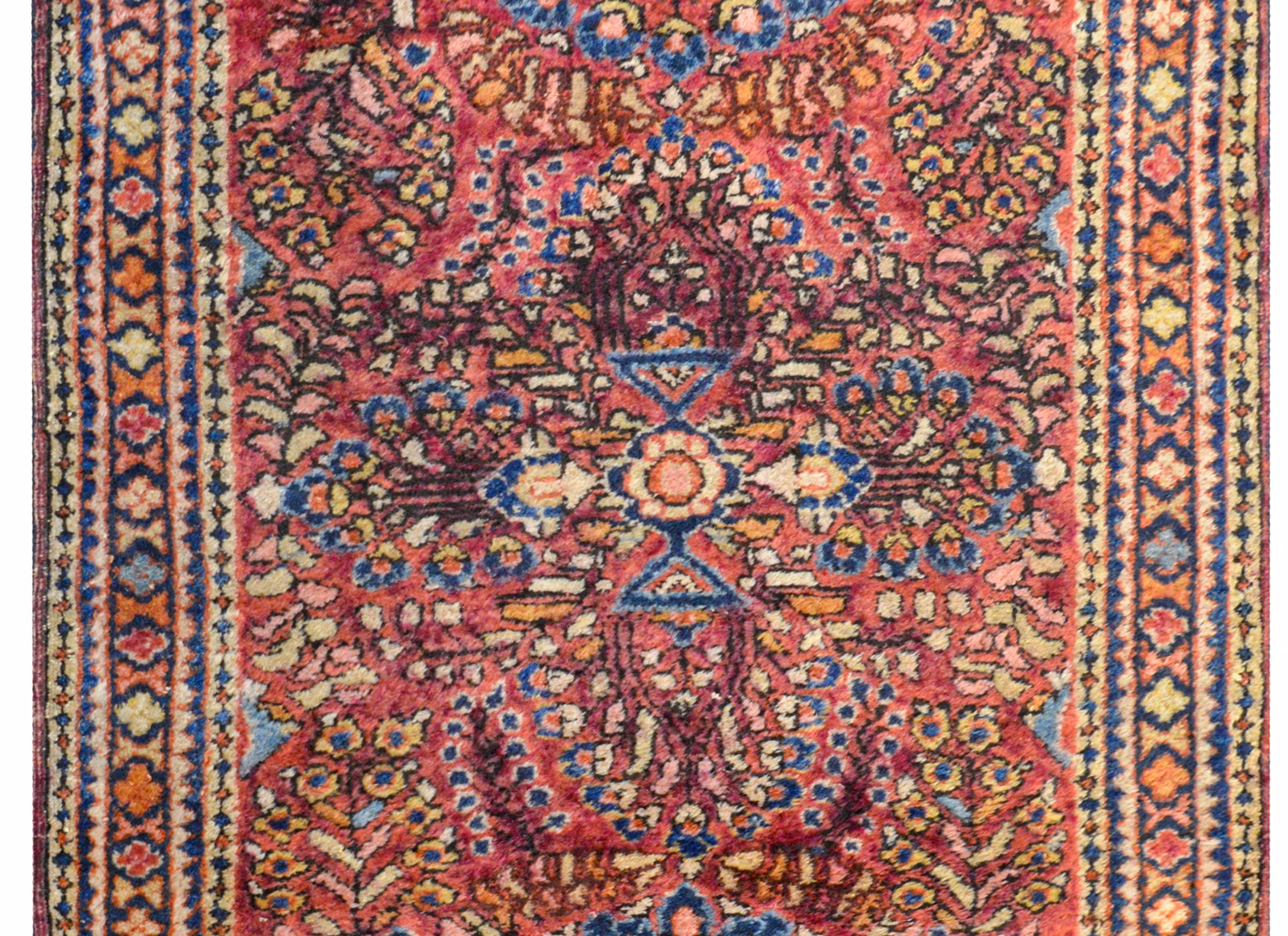 A traditional early 20th century Persian Sarouk rug with a large-scale mirrored floral tree of life pattern woven in cranberry, gold, indigo, and orange, on a cranberry background. The border is composed with multiple petite geometric pattered