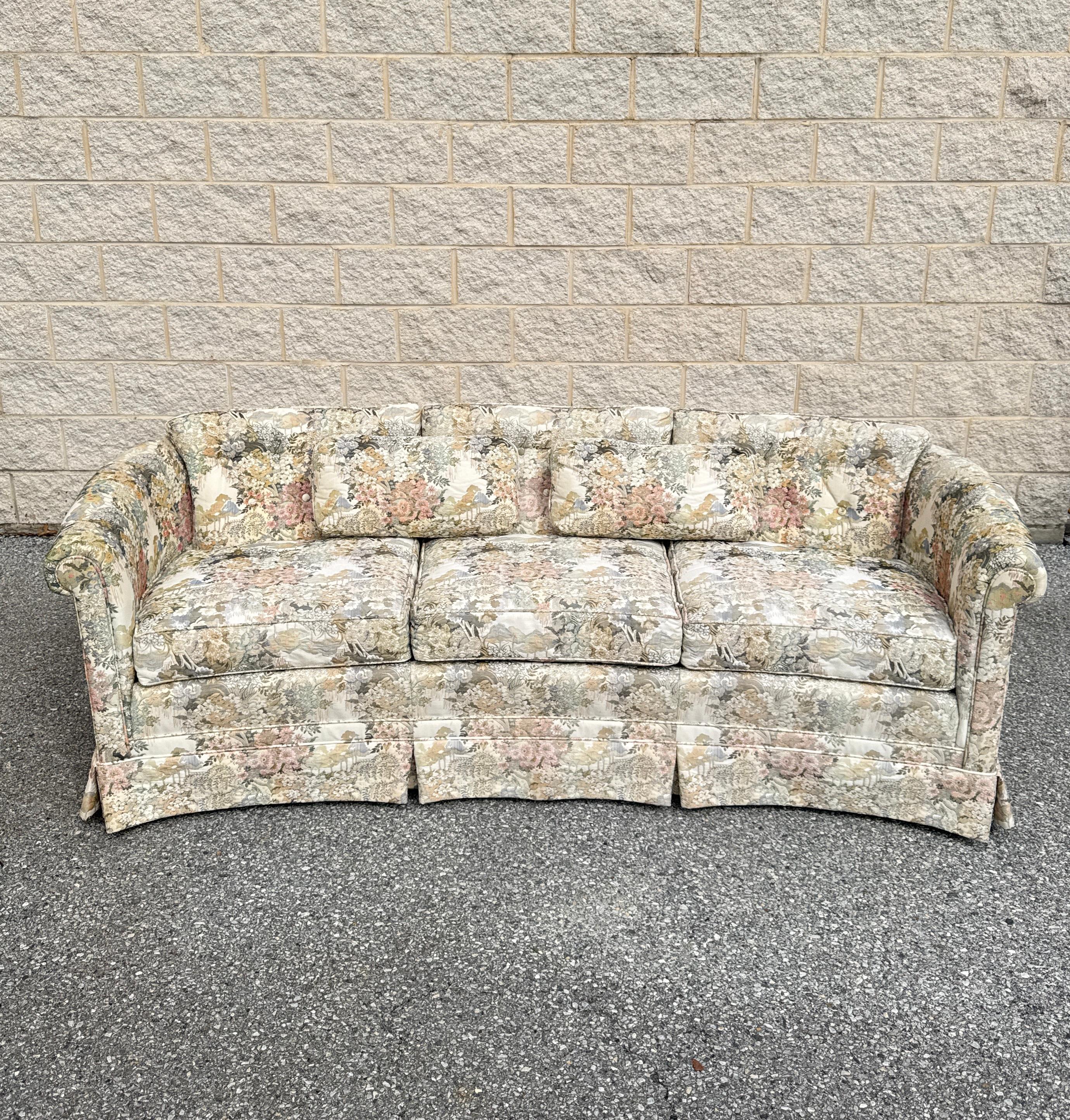 Late 20th-century traditional curved sofa featuring pastel floral upholstery with a Chinoiserie-inspired pattern. Designed with rolled arms, a skirted base, and three-seat cushions. The gently curved frame provides a refined silhouette suitable for