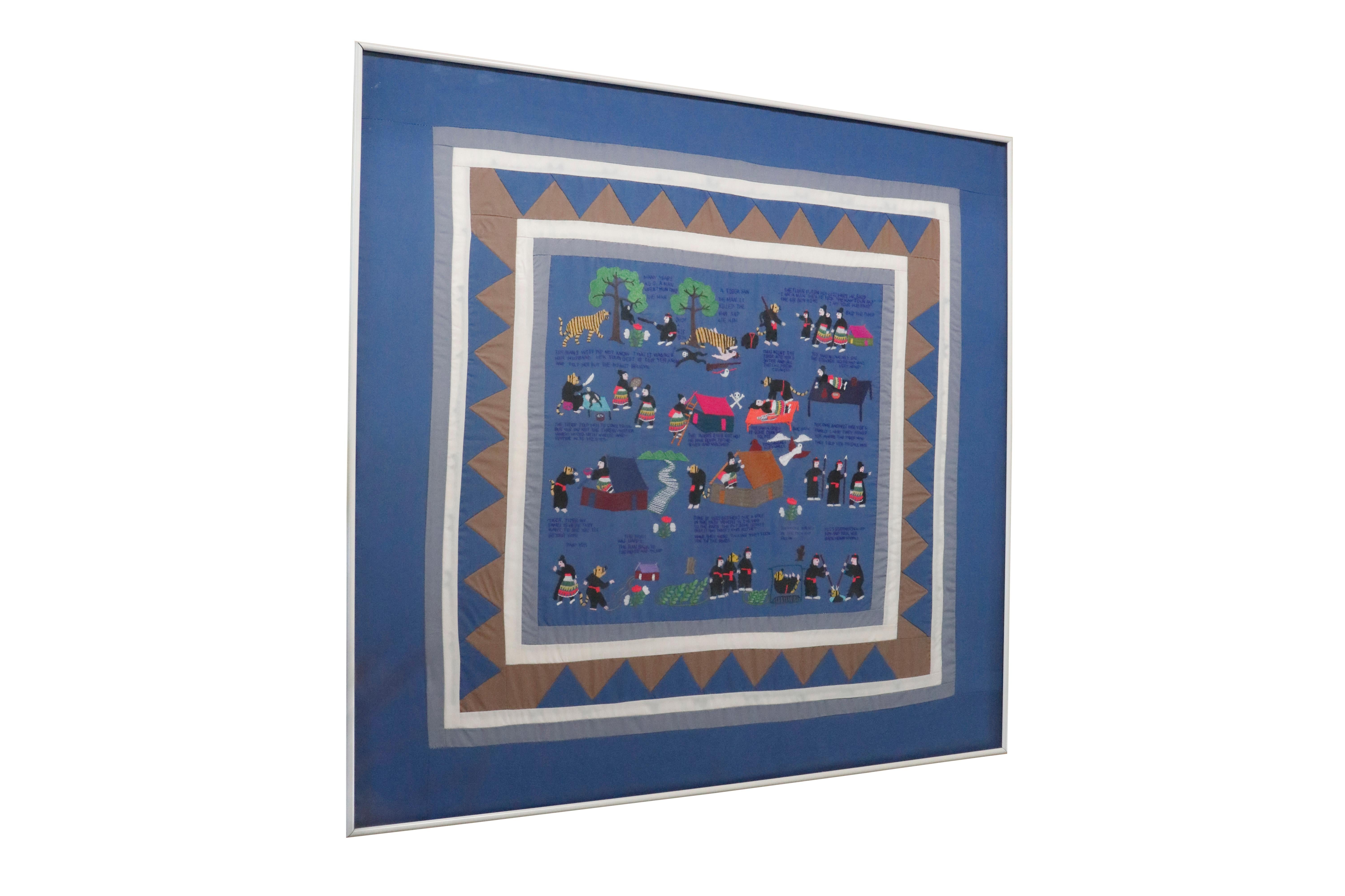 A traditional story quilt. A blue background is embroidered to tell the story of a tiger who kills and impersonates man who shoots a gibbon. Framed with a light blue and white border decorated with brown and blue triangles. Set behind glass in a