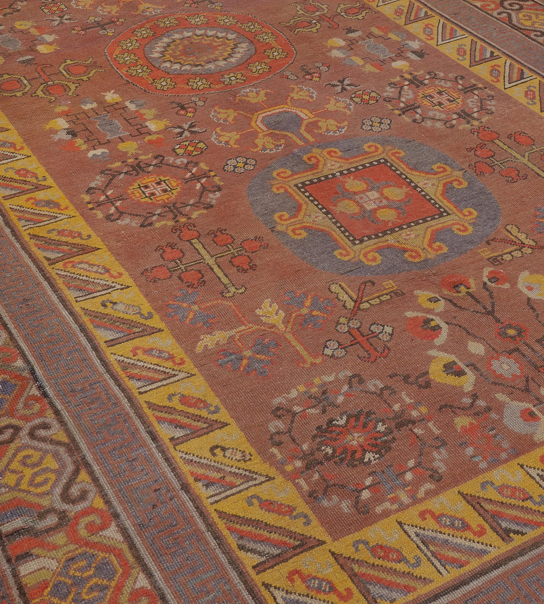 This traditional hand-woven Samarkand Khotan rug has a rust field with a profusion of auspicious symbols and pomegranate motif enclosing a triple column of roundel medallions, in a profusion of tradition polychrome Chinese borders.