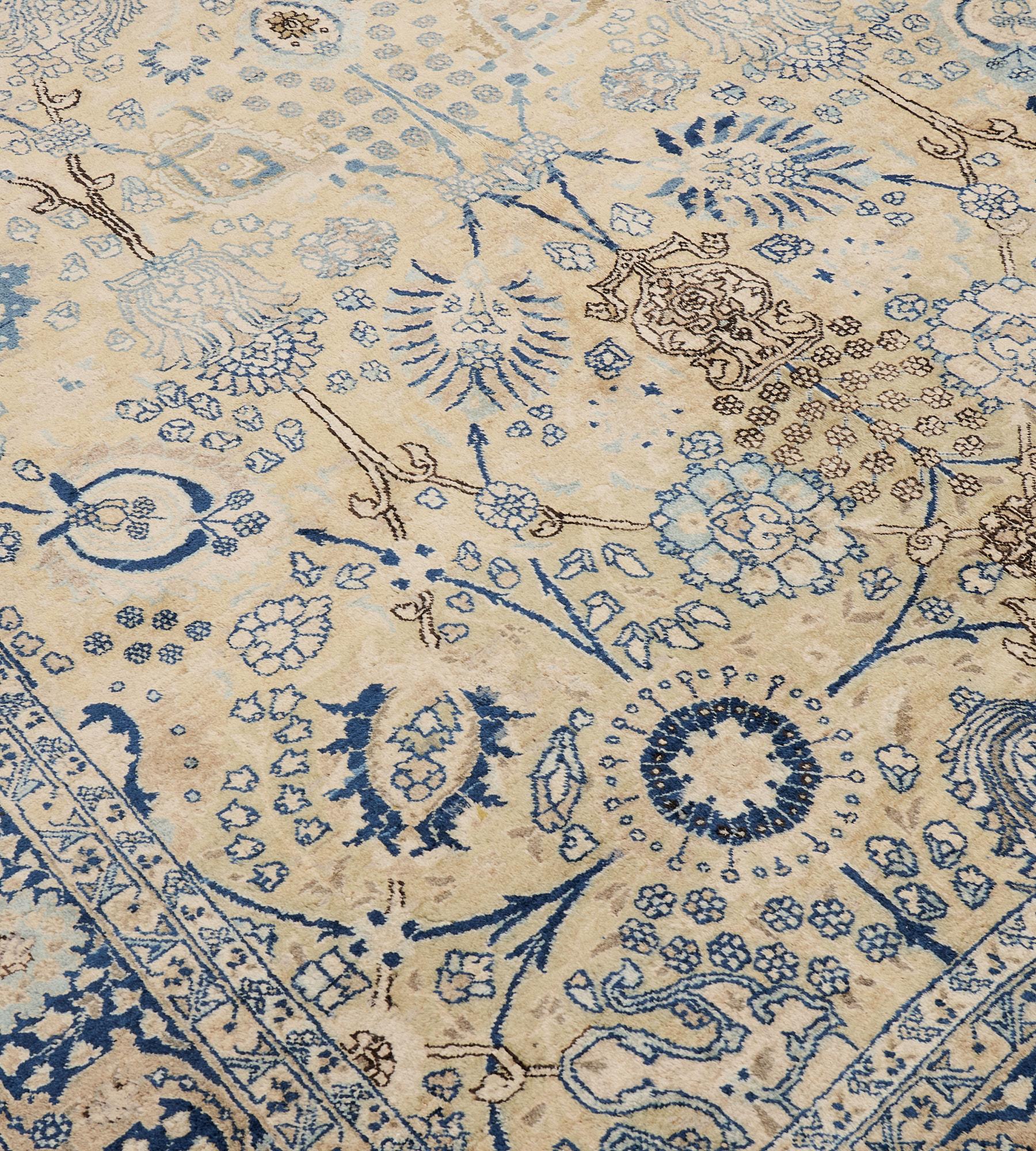 Ce tapis traditionnel persan Tabriz tissé à la main présente un champ brun chamois doux avec une colonne centrale de vases fleuris noir charbon, bleu indigo, bleu clair et ivoire reliés par des palmettes et des lianes florales, dans une bordure bleu