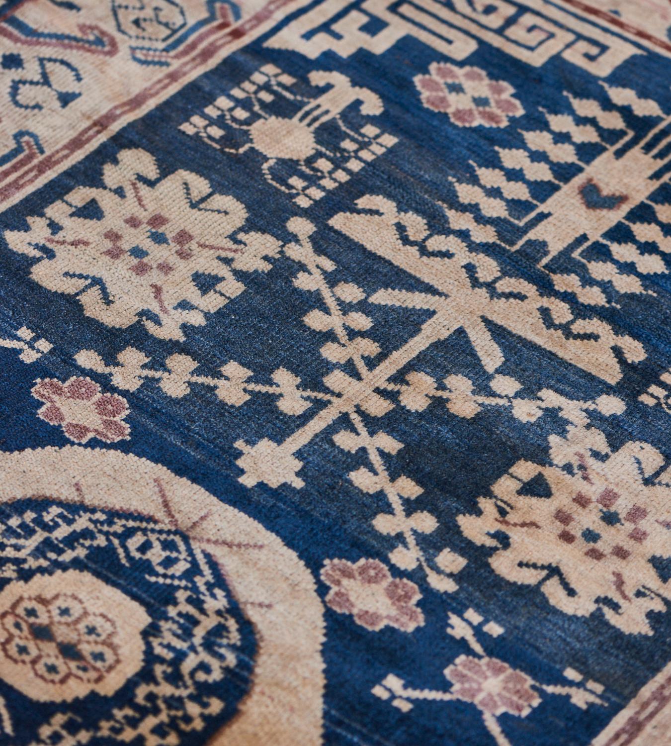This traditional Khotan rug has a shaded navy blue field scattered with floral sprays, hooked floral motifs, and other auspicious designs around central beige roundel containing a flowing vine around a stellar flowerhead, there are stepped angular