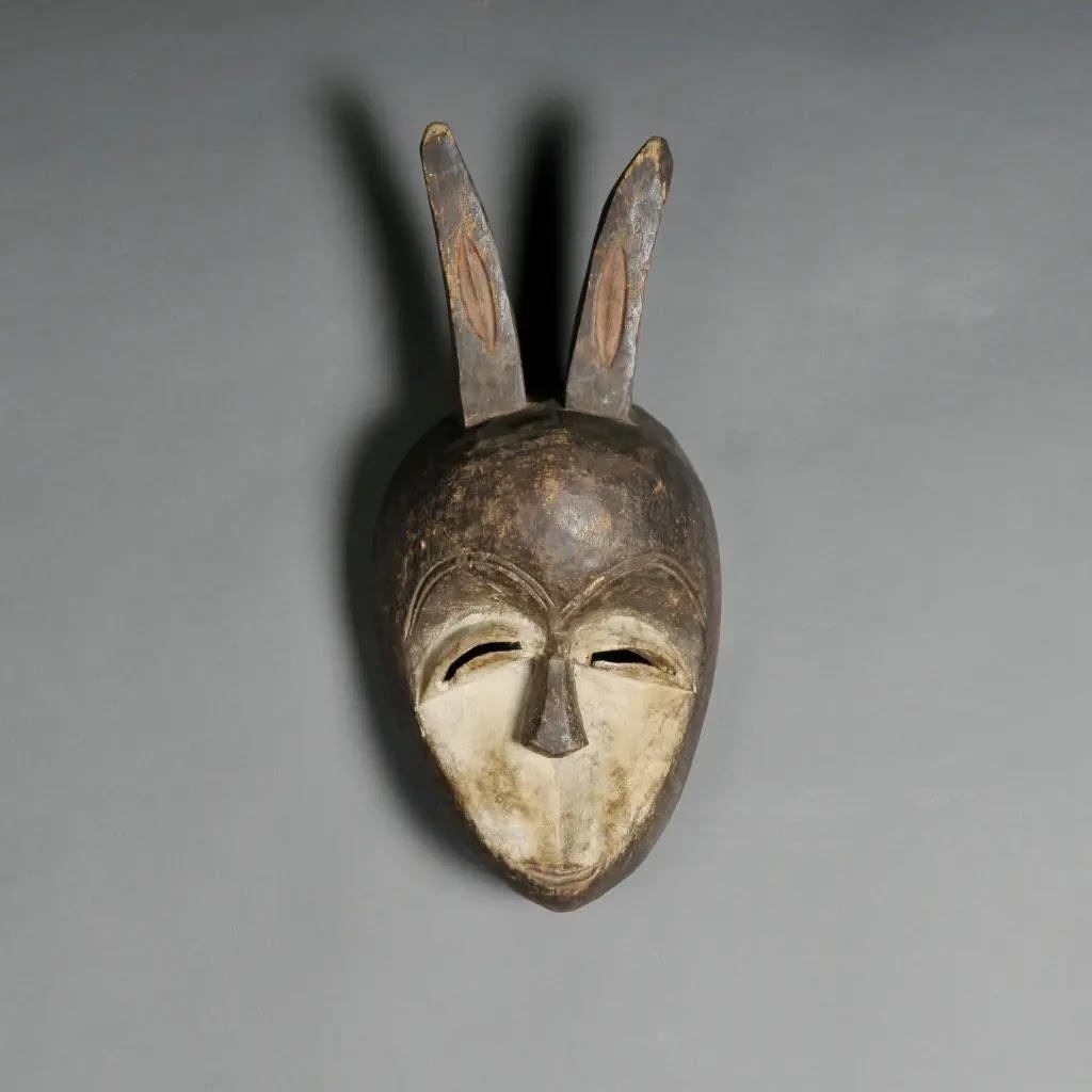 Diese traditionelle afrikanische Kéwélé-Maske aus dem Kongo ist ein beeindruckendes ethnisches Kunstobjekt, das aus gealtertem Holz handgeschnitzt und mit einer sorgfältig aufgetragenen natürlichen Patina versehen wurde. Ihre längliche Form und die