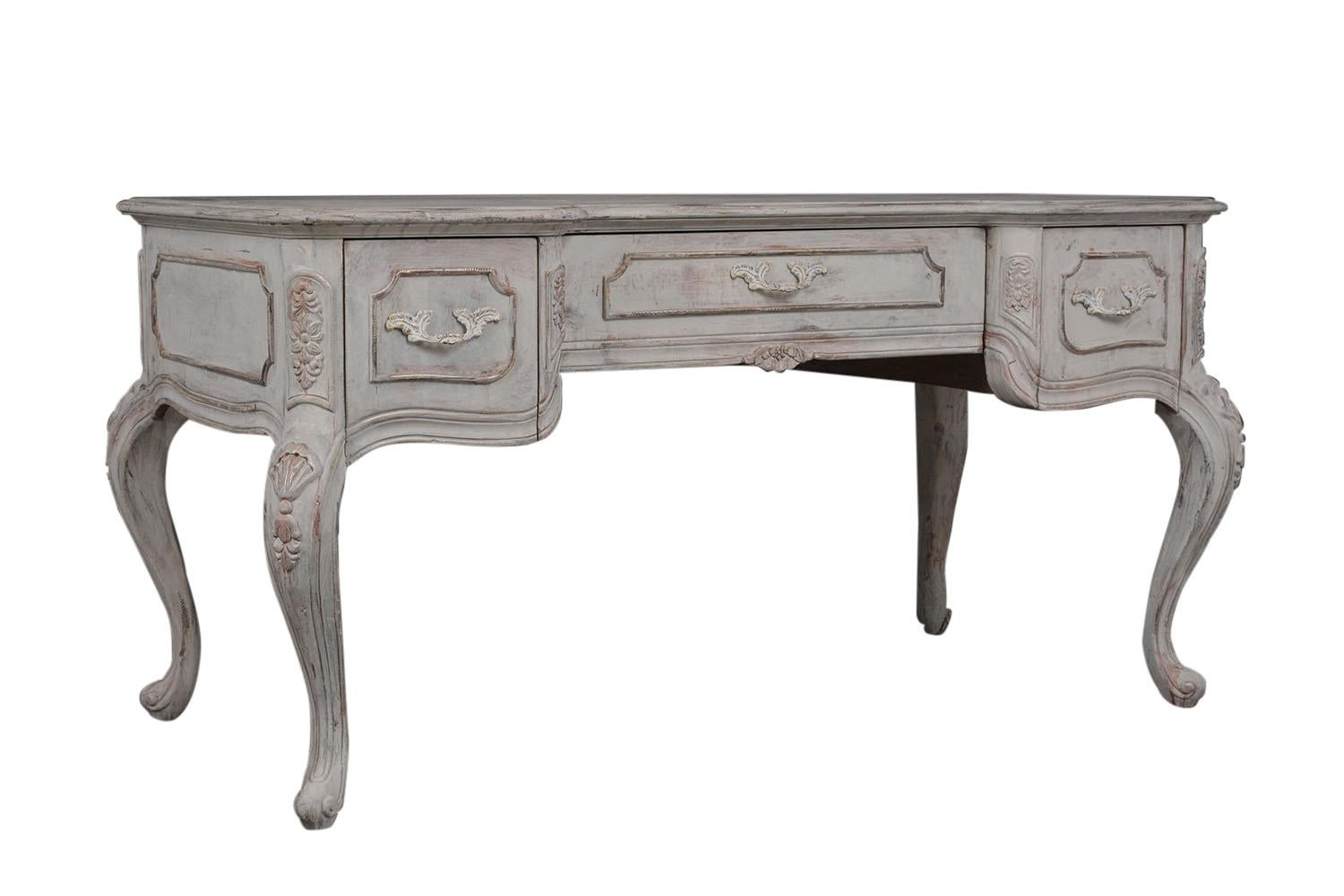 This Vintage French Louis XV Desk is made out of solid wood and has been newly painted in an off gray color with a beautiful hand-done distressed finish. The desk features a solid beveled wooden top, two side drawers & large center drawer front