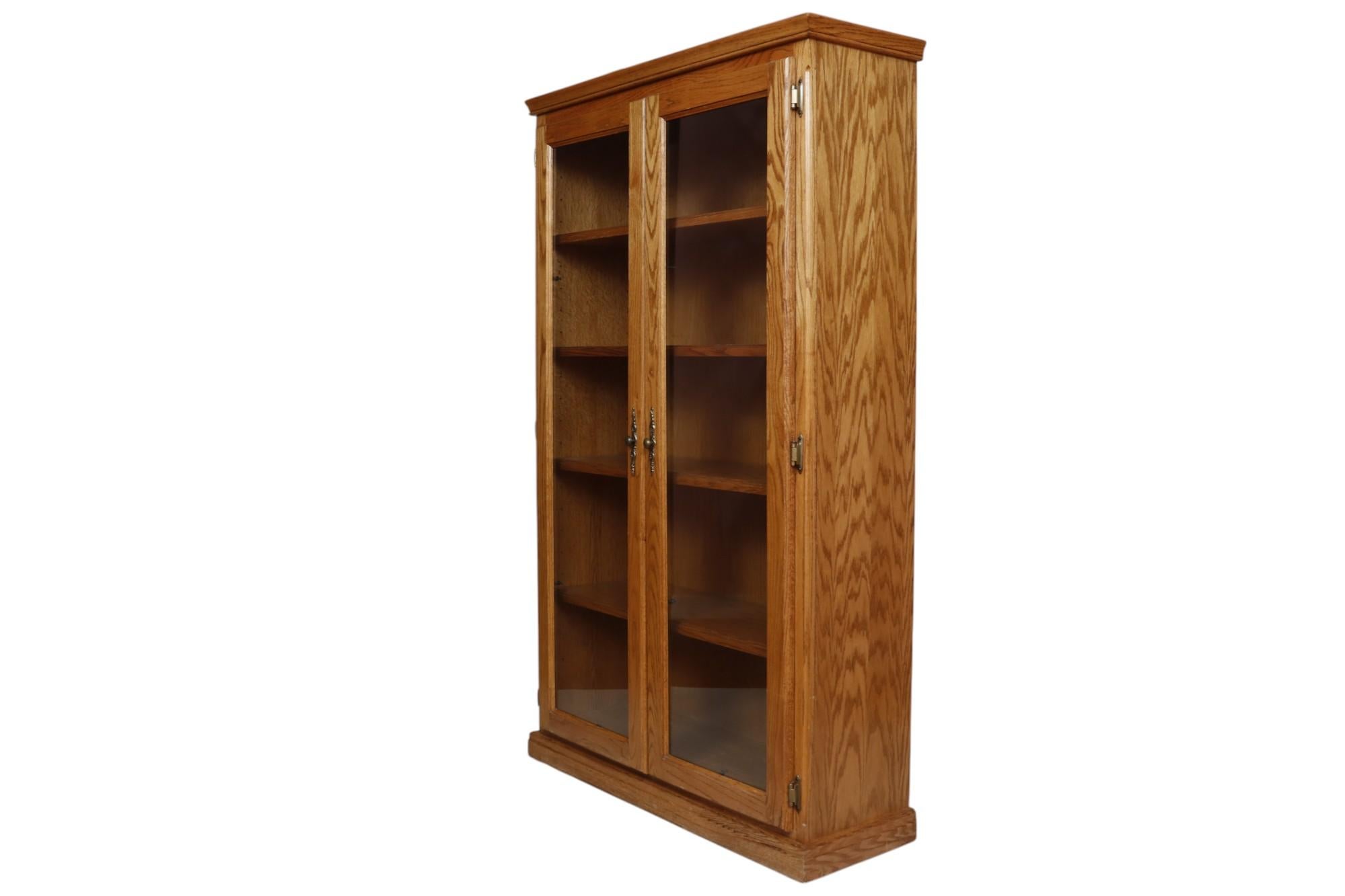 A traditional wooden bookcase made of oak with oak veneers. The solid oak frame supports four oak veneered shelves behind two glass doors. The doors open with round brass mushroom knob handles with long pressed backplates. 