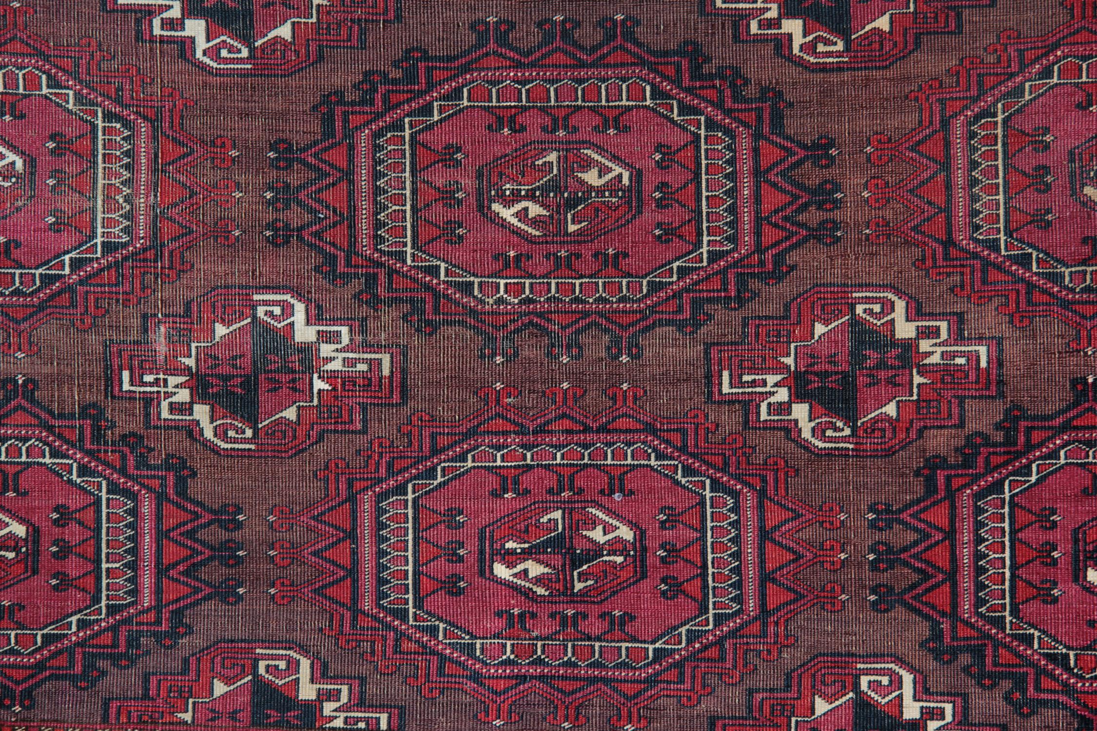 Esta alfombra antigua tradicional es un claro ejemplo de las alfombras tejidas en la década de 1880 y presenta un fondo rojo con motivos geométricos tejidos en colores beige y marrón en contraste sobre un fondo rojo intenso. El diseño central de