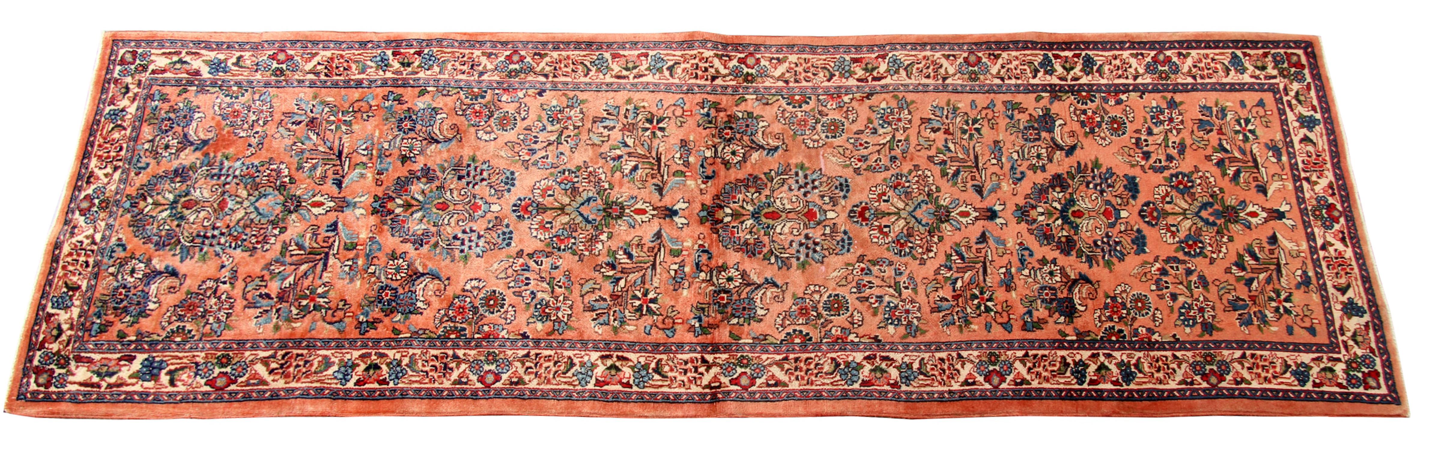This elegant rust vintage rug has been woven on a pink-orange background with a highly decorative repeat pattern border featuring blue, brown, and red accents. An intricate floral border then encloses this. Both the colour palette and sophisticated