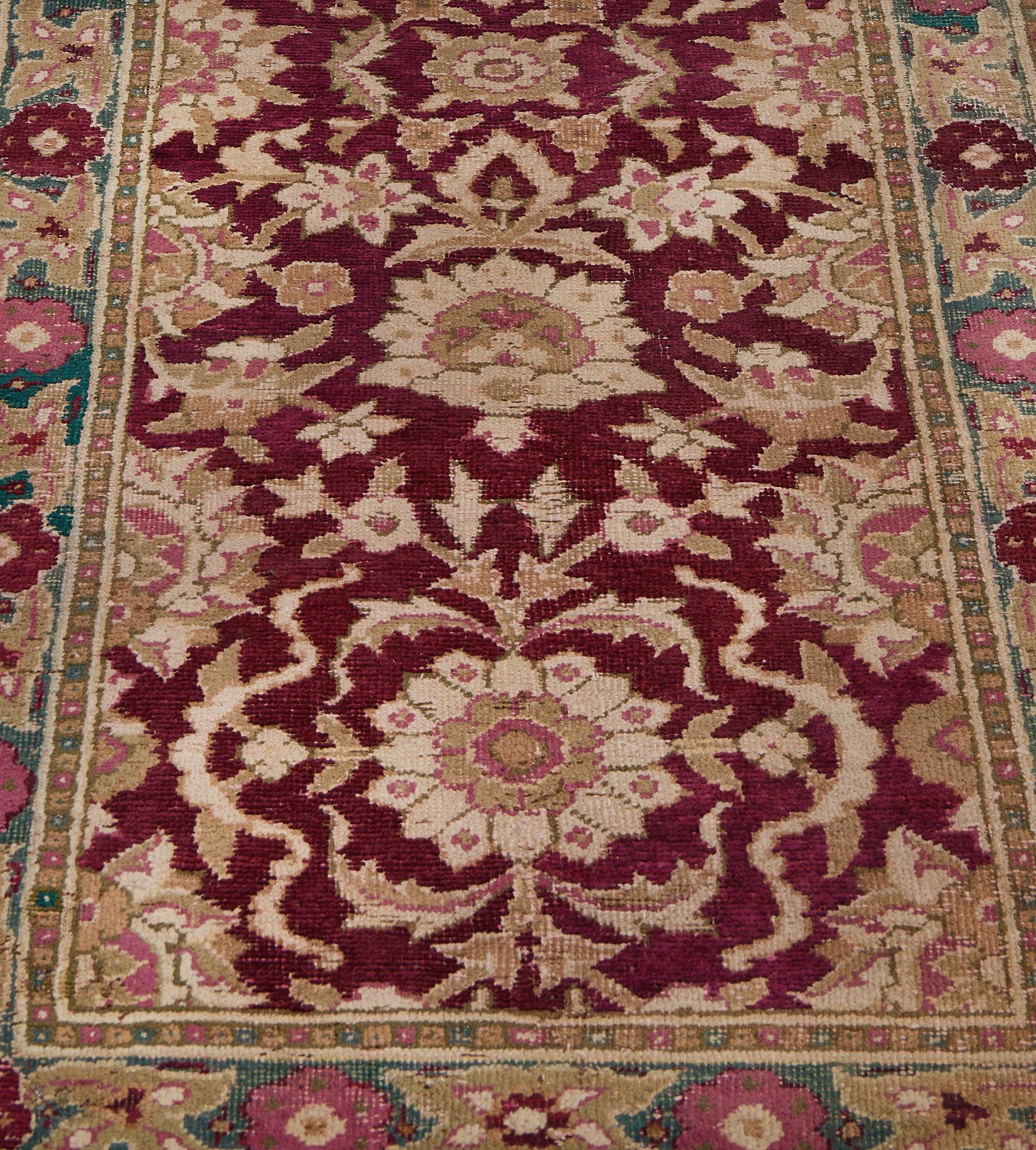 This antique Indian Agra runner has a burgundy-red field with a column of ivory, pink and sandy-brown rosettes and palmettes surrounded and linked by scrolling floral and leafy vine, similar part palmettes at each side, in a sea-blue border of