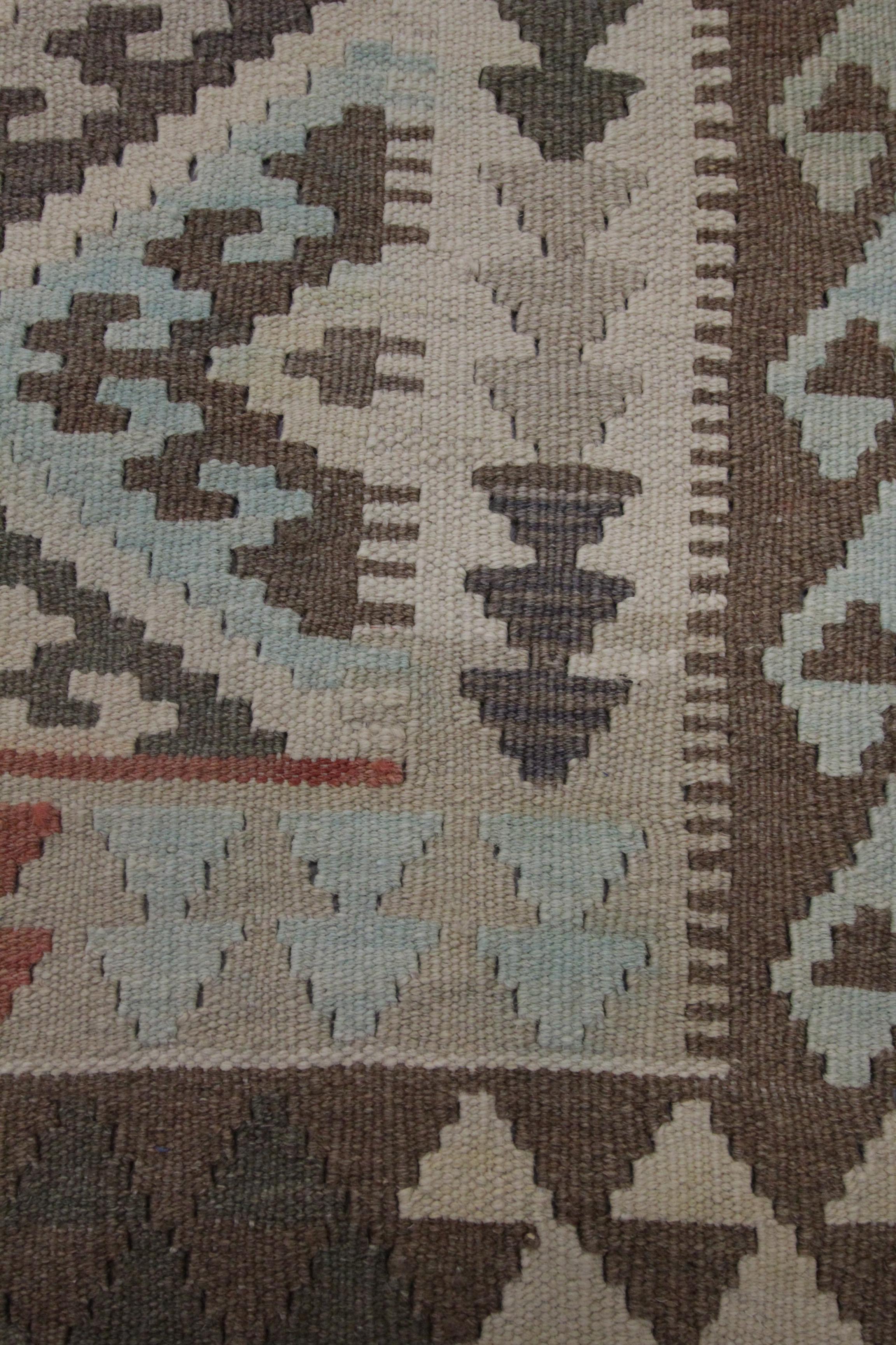 This bold traditional wool Kilim rug is a handwoven Afghan Kilim constructed in the early 21st century, circa 2010-15. The design features a repeating motif pattern made up of hook medallions in blue, beige, and brown accents. A layered border then