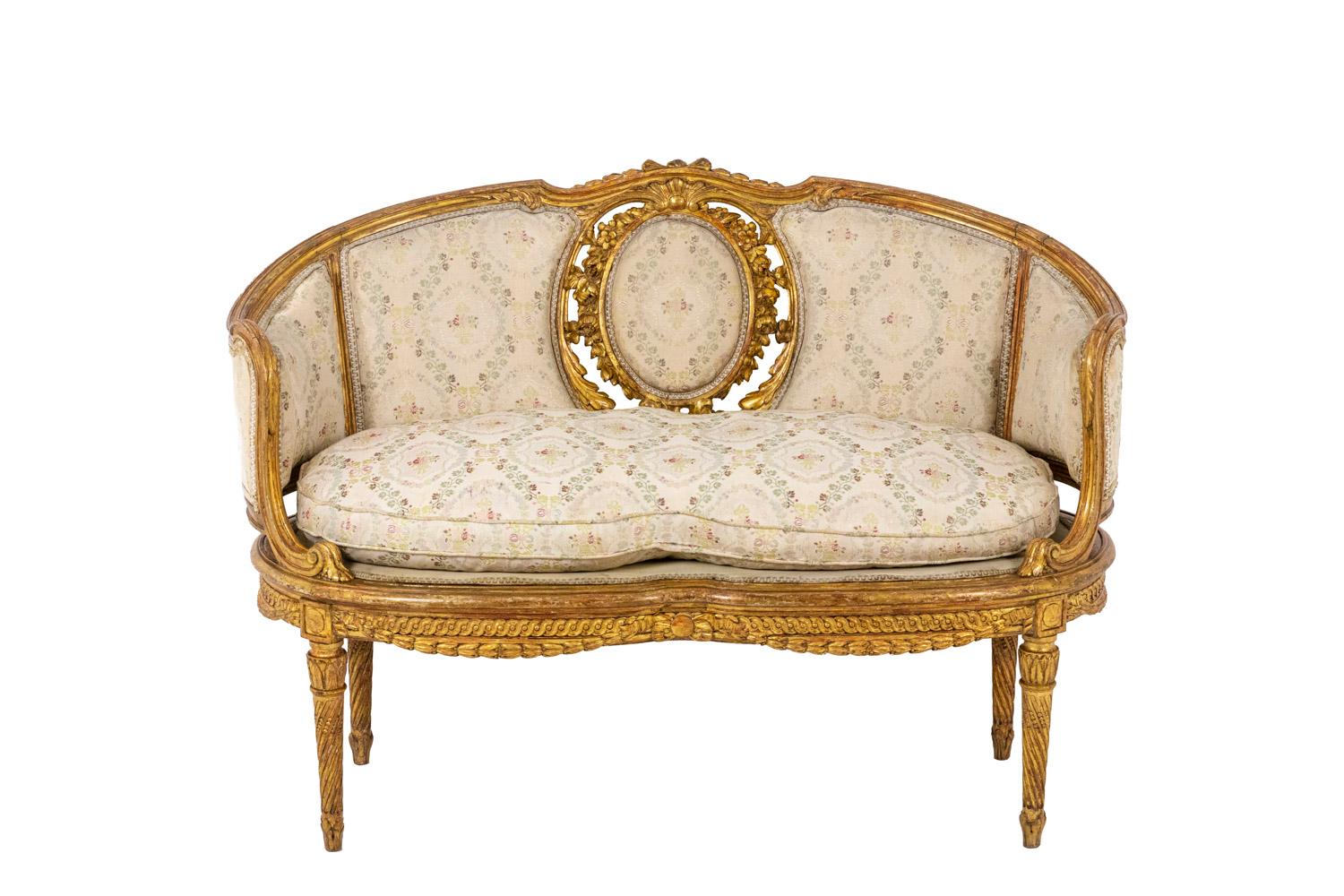Transition style two places sofa in molded and gilt wood standing on four straight tapered and fluted legs. The belt is adorned with garlands and a frieze of droplets. Arm supports slightly behind the leg line. Armrests with ears. Curved back with a