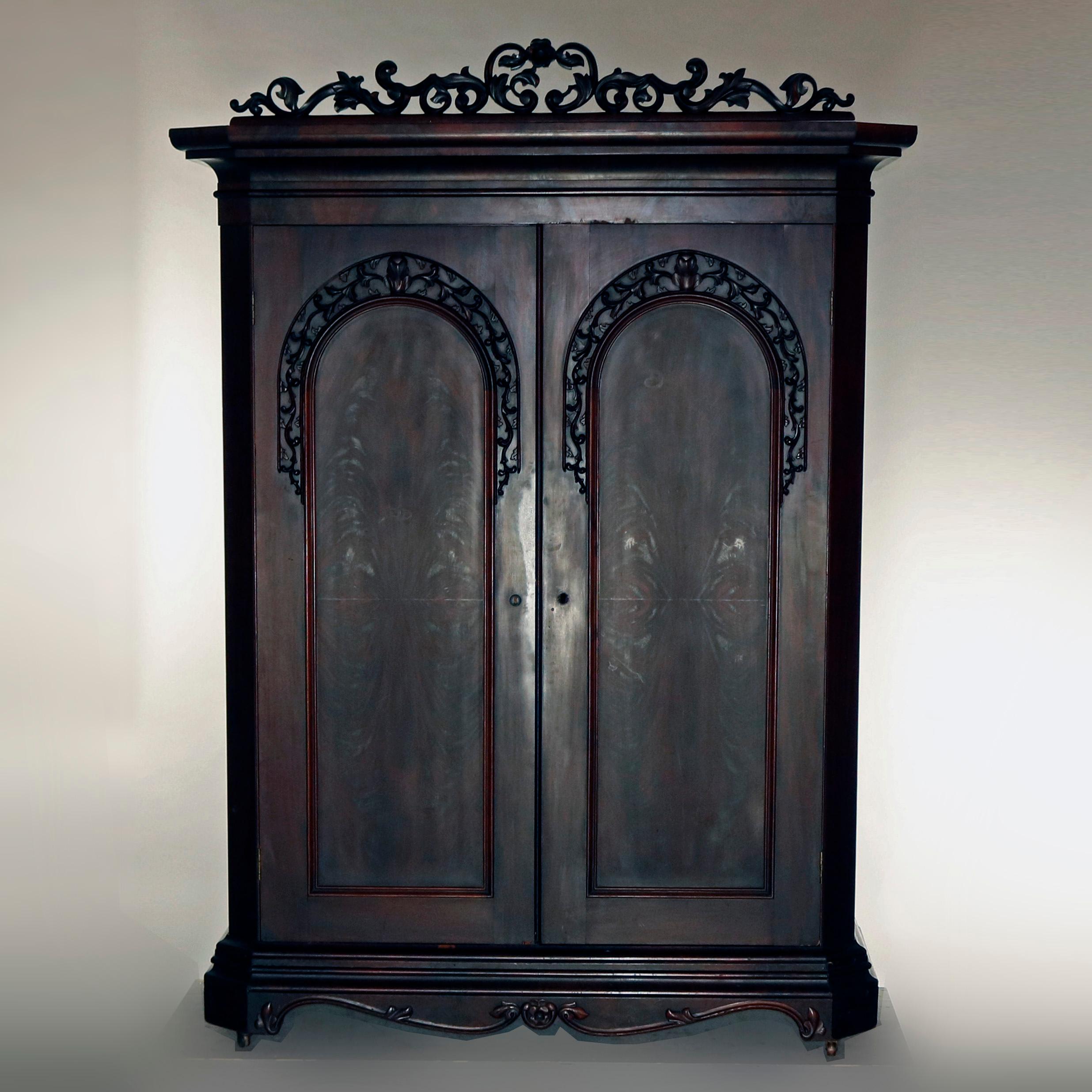 An antique transitional Empire into Victorian wardrobe features flame mahogany construction in Empire form and incorporating elements of Victorian including the carved and pierced scroll and foliate crest, arch panelled double doors with applied