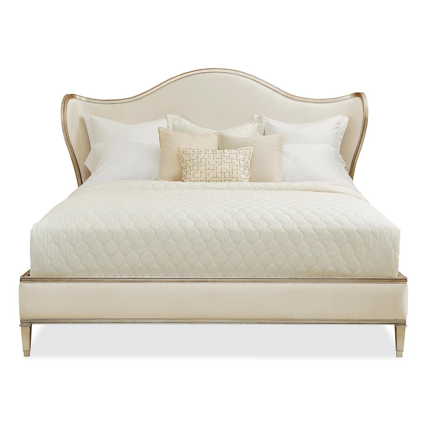 A Transitional Style Upholstered King Bed with a serpentine headboard and curved wings. The exposed carved wood rail is finished in radiant Auric silver leaf. Fully upholstered in a cream fabric. This piece has a low footboard and square-tapered