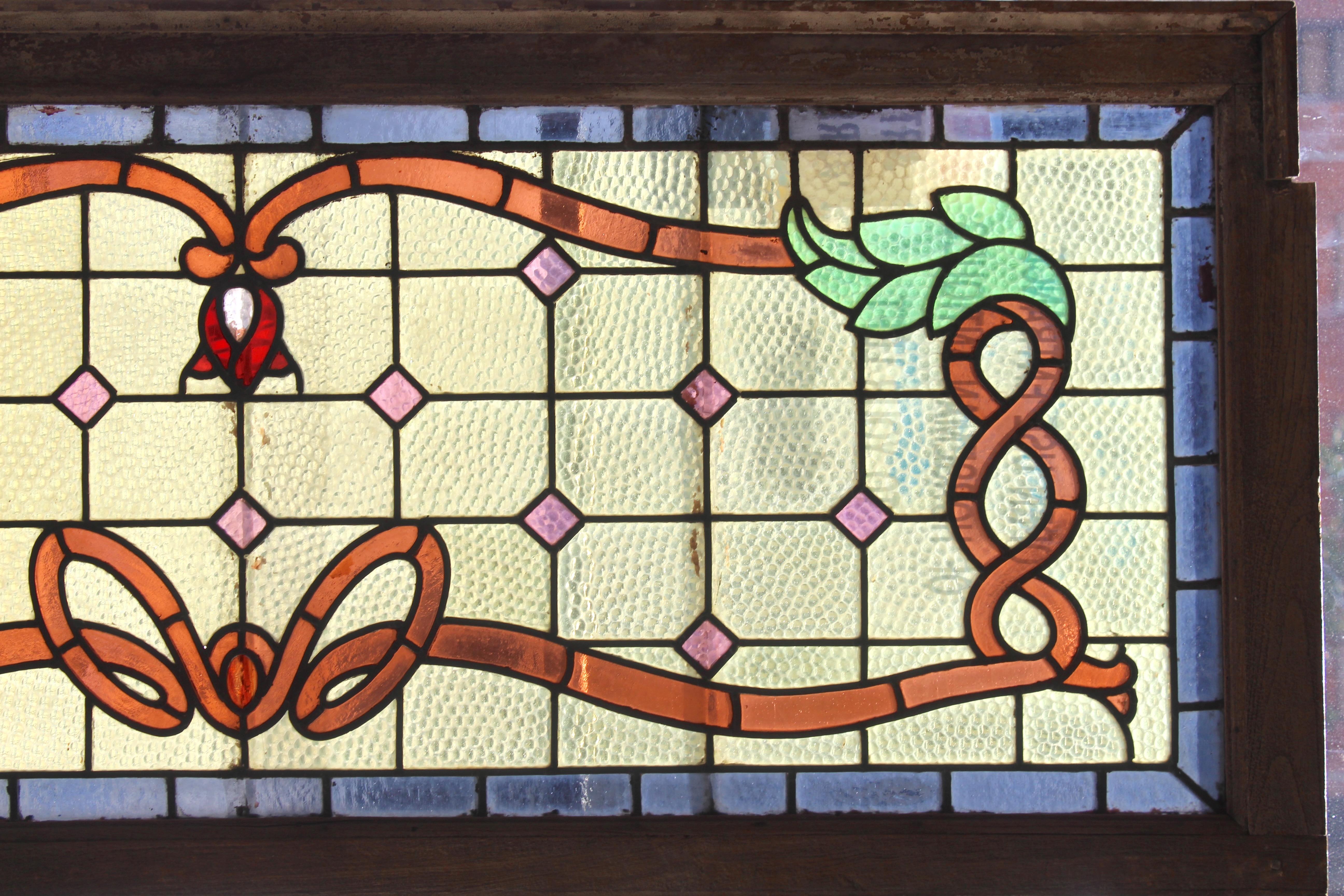Long transom window with elaborate stained glass designs. Textured glass, scrolling ribbons, pink diamonds, and a deep blue trim around.
Currently housed in a wooden frame - the overall dimensions are for the stained glass and the frame.
Located in