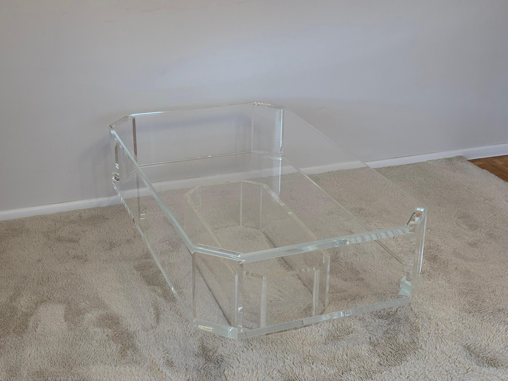 Sleek modern clear acrylic coffee table featuring a faceted architectural silhouette with an inset plinth-style base. Crafted from thick, high-clarity cast acrylic, the table appears visually weightless while offering strong structural presence. The