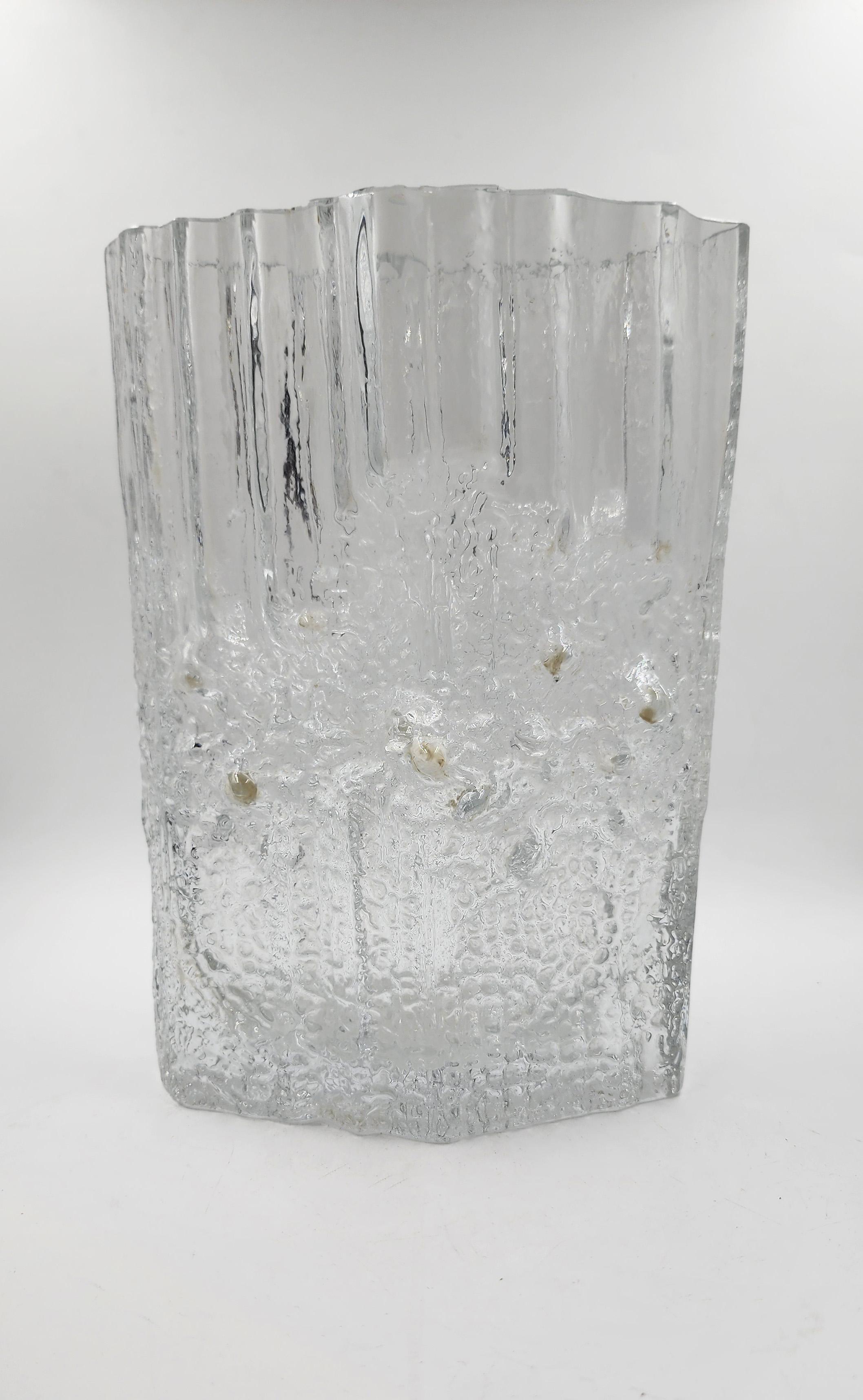 Jarrón de cristal finlandés transparente de Tapio Wirkkala
Este jarrón de cristal finlandés, diseñado por Tapio Wirkkala, es una pieza transparente de forma rectangular irregular. Su superficie exterior presenta una textura que simula hielo