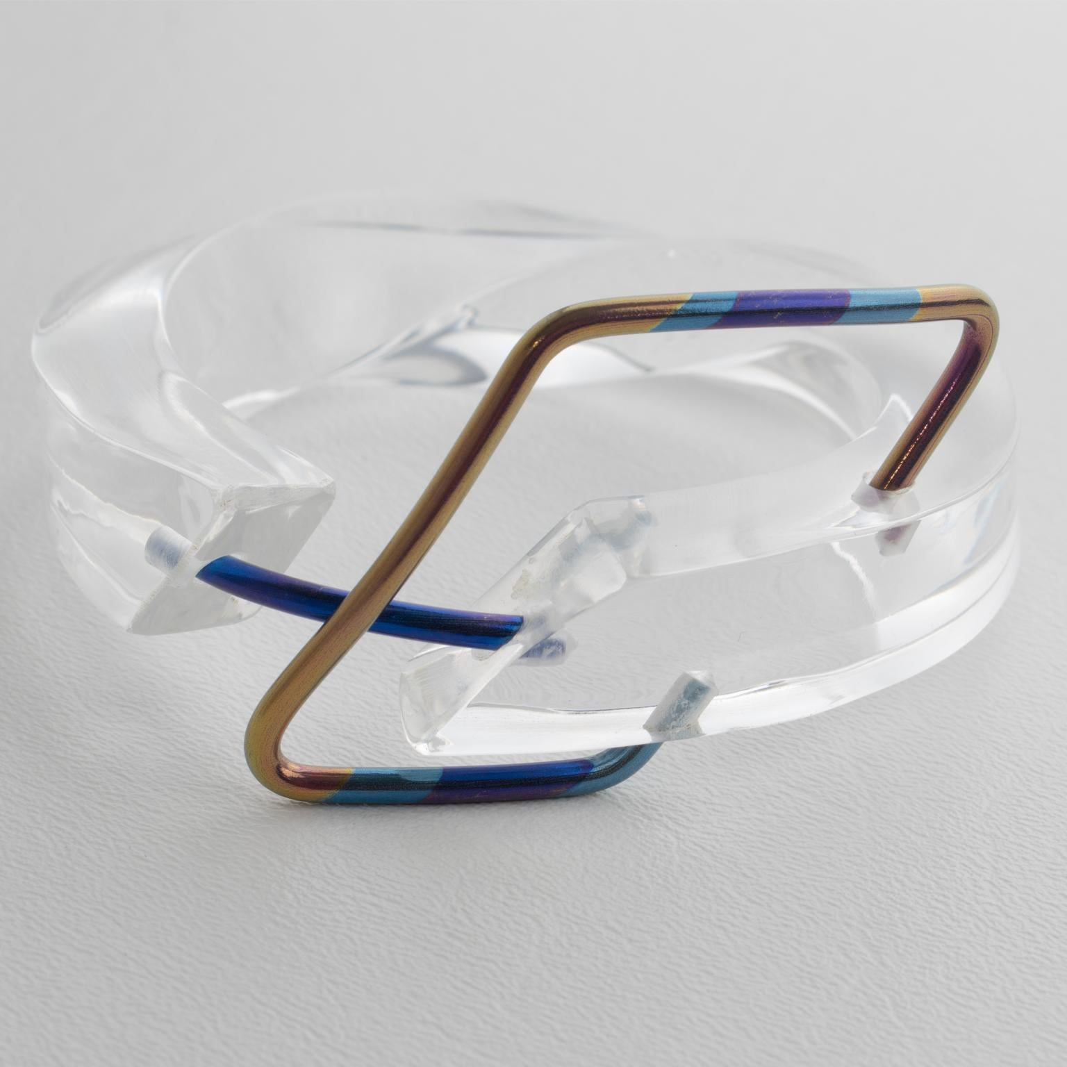 This stunning Space Age sculptural Lucite bracelet bangle features a chunky hand-crafted, open twisted thick band shape in transparent color, adorned with an anodized aluminum belt buckle decor. The belt buckle features luminous cerulean, cobalt,