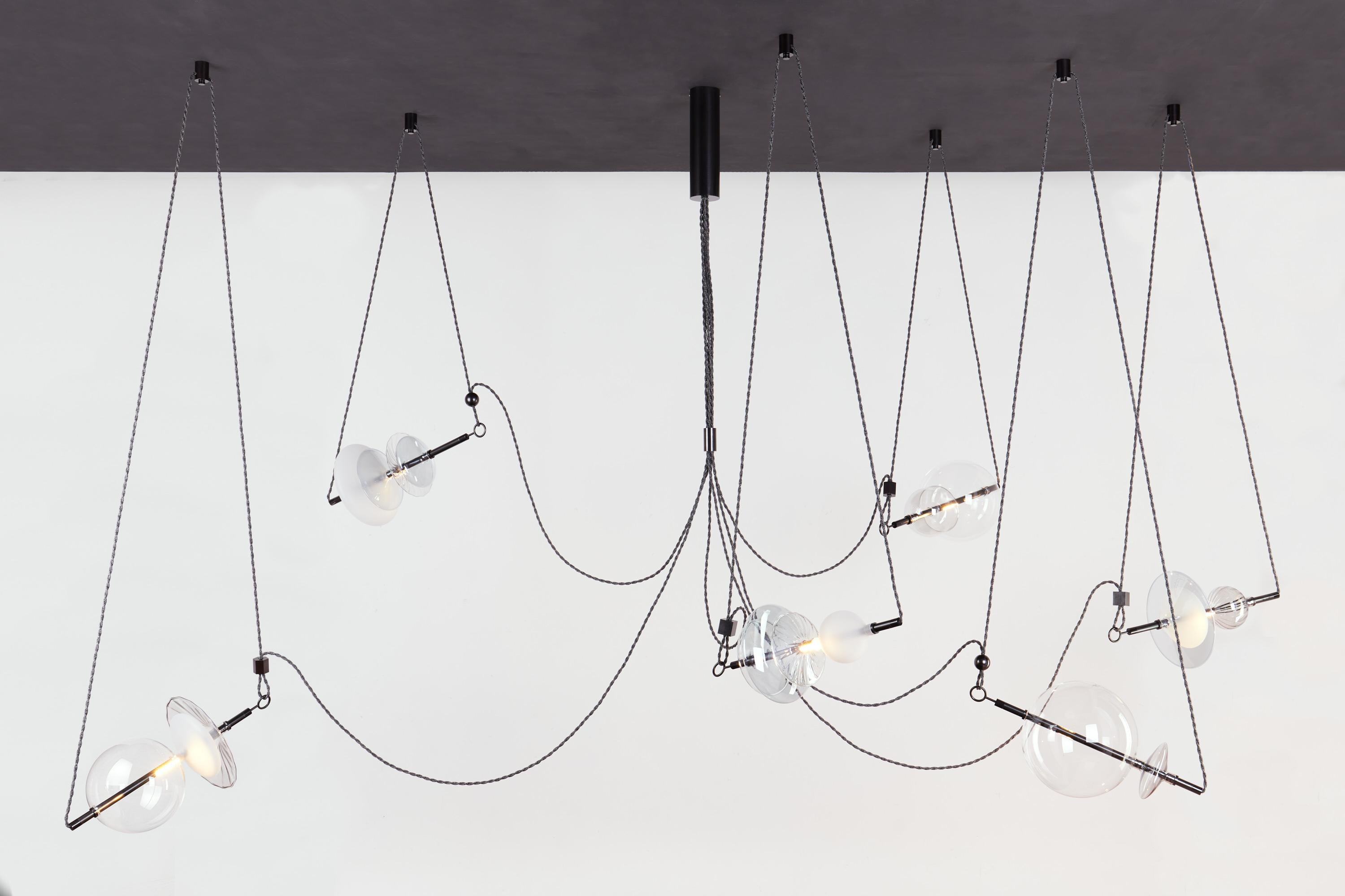 The Trapezi Black contemporary chandelier is inspired by the idea of a Circus Trapeze Artist.

Hand blown glass in a variety of forms and colors is combined with brass bars and hung by Linen Cords from hub units on the ceiling.
The glass is left