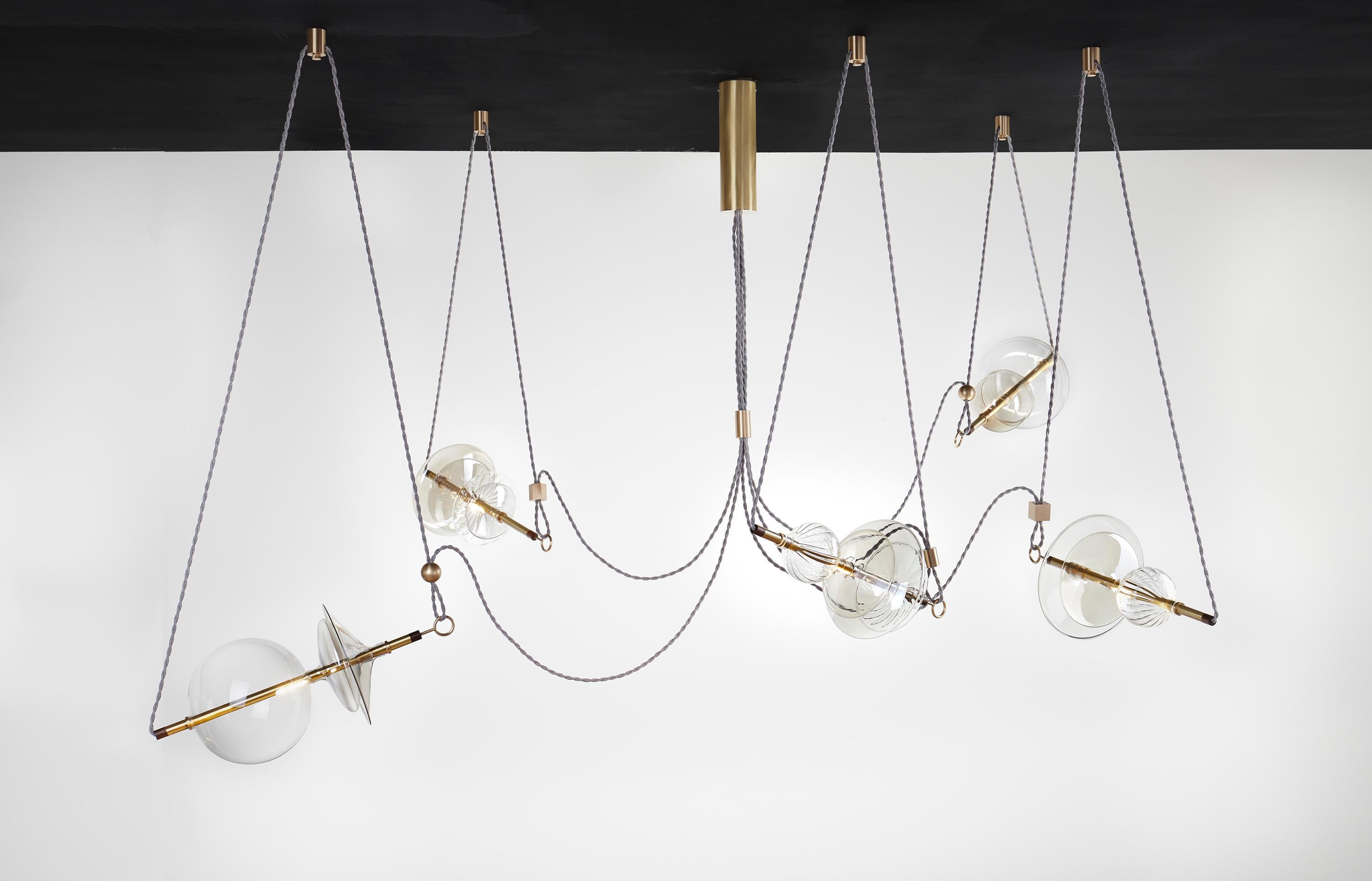 The Trapezi contemporary chandelier is inspired by the idea of a Circus Trapeze artist.

Hand blown glass in a variety of forms and colors is combined with brass bars and hung by Linen Cords from hub units on the ceiling.
The glass is left