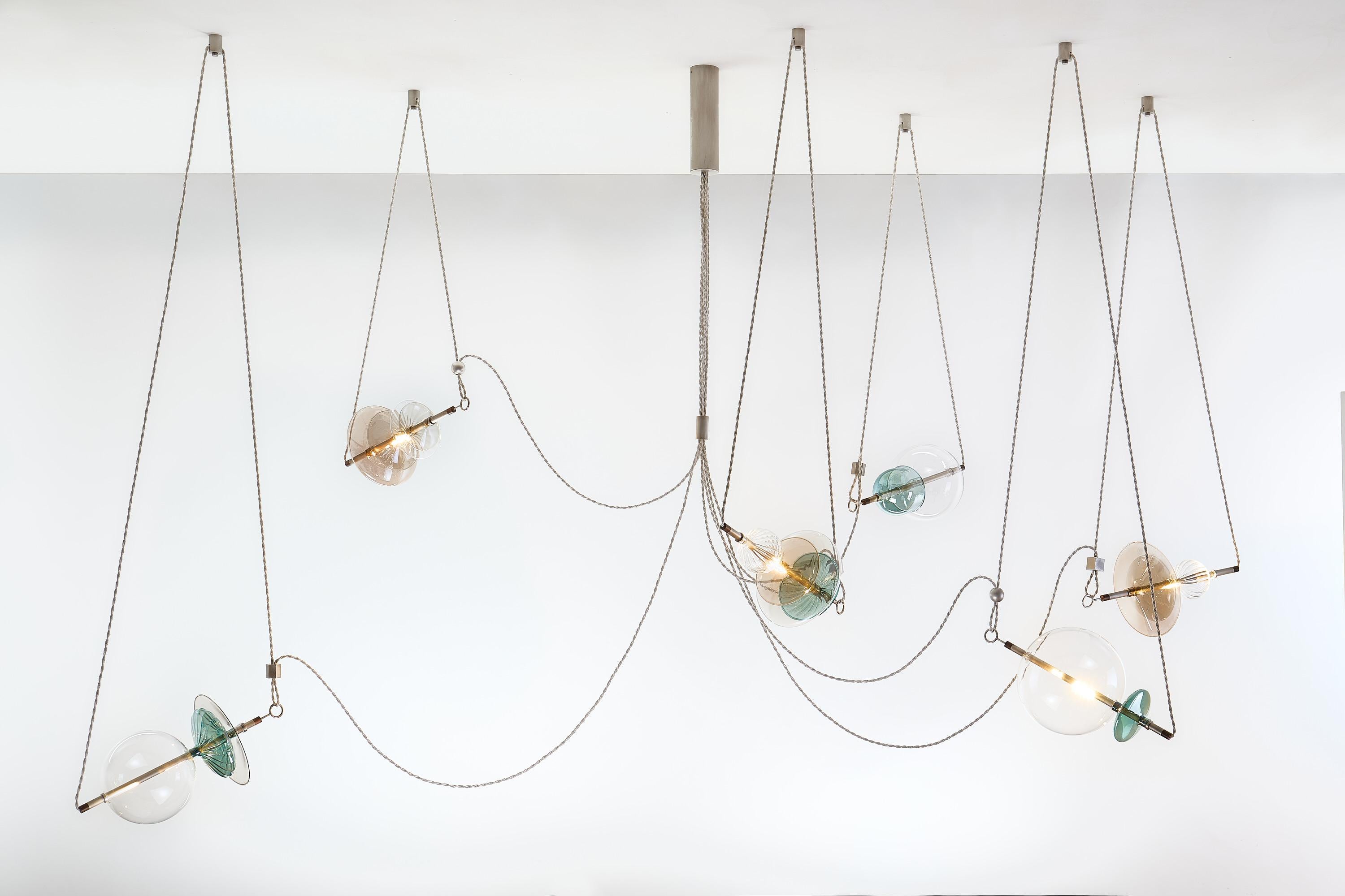 The idea of a Circus Trapeze Artist inspires the Trapezi contemporary chandelier.

Hand-blown glass in a variety of forms and shades is combined with brass bars and hung by Linen Cords from hub units on the ceiling.
The glass is clear or