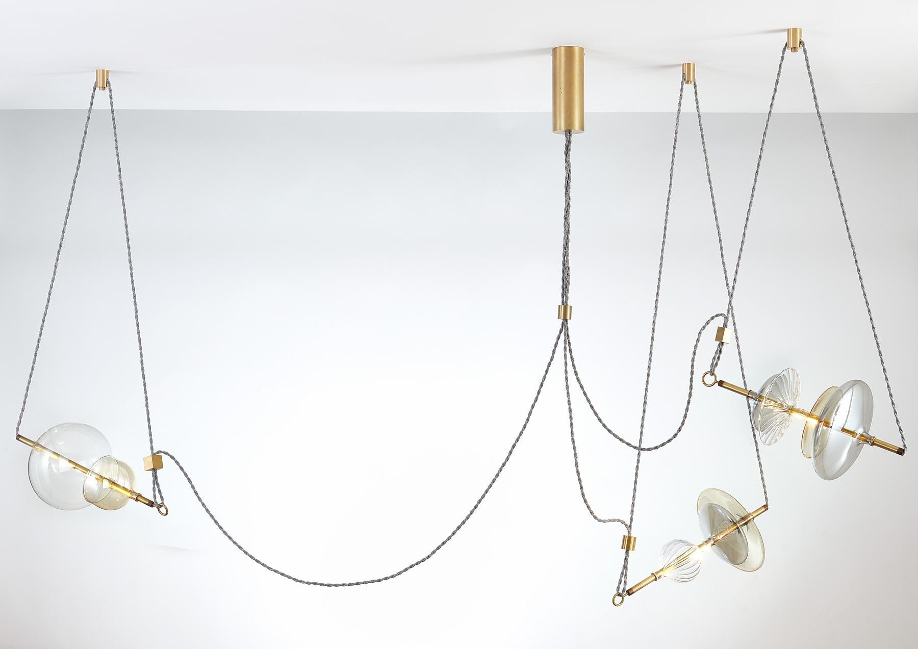 The Trapezi contemporary chandelier is inspired by the IDEA of a Circus Trapeze Artist.

Hand blown glass in a variety of forms and colors is combined with brass bars and hung by Linen Cords from hub units on the ceiling.
The glass is left