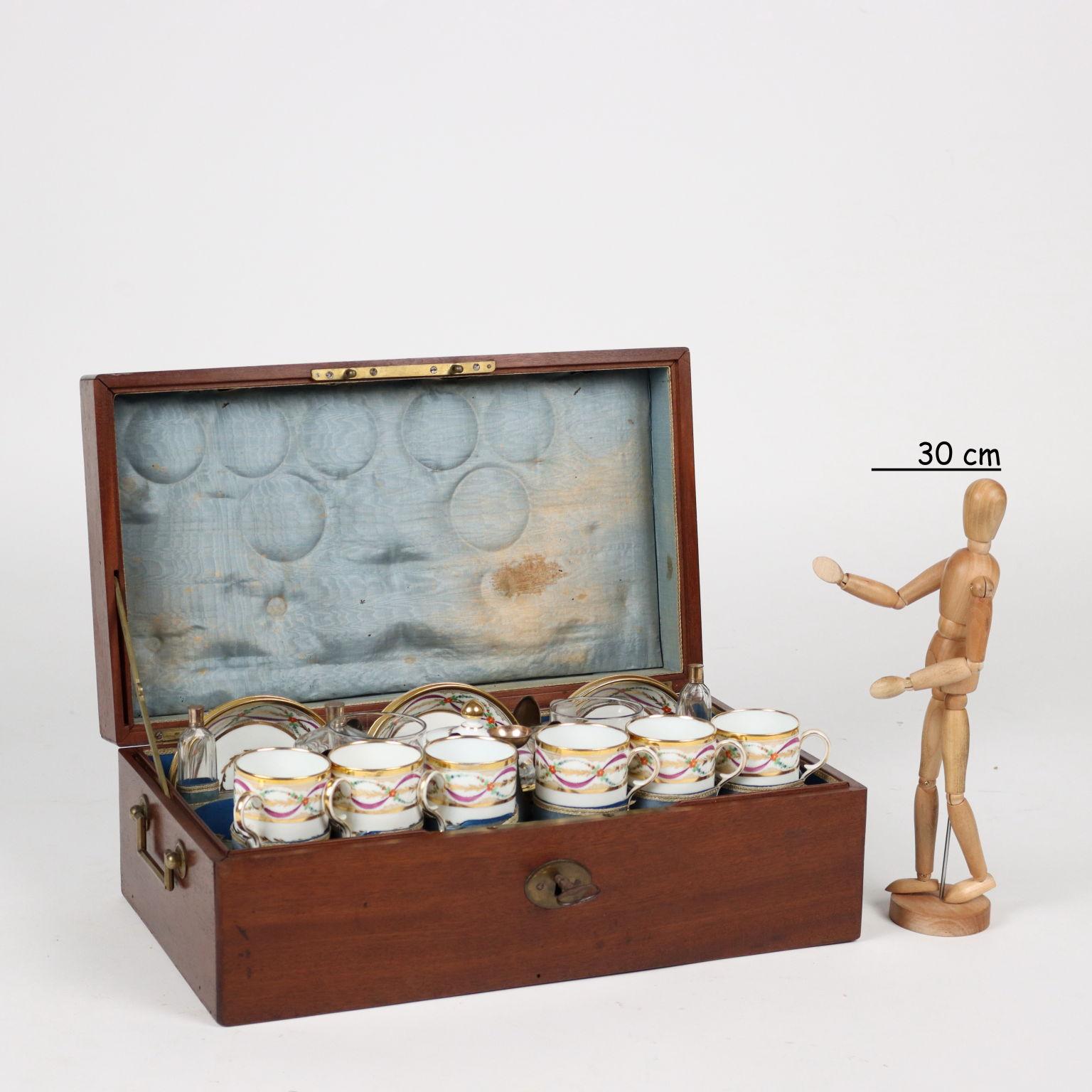 Baule in legno di mogano con prese in ottone. All'interno, un set in porcellana francese finemente decorato in oro zecchino e policromo con nastri e ghirlande di fiori. Servizio composto da sei tazze da tè con piattini e zuccheriera. Mancano la