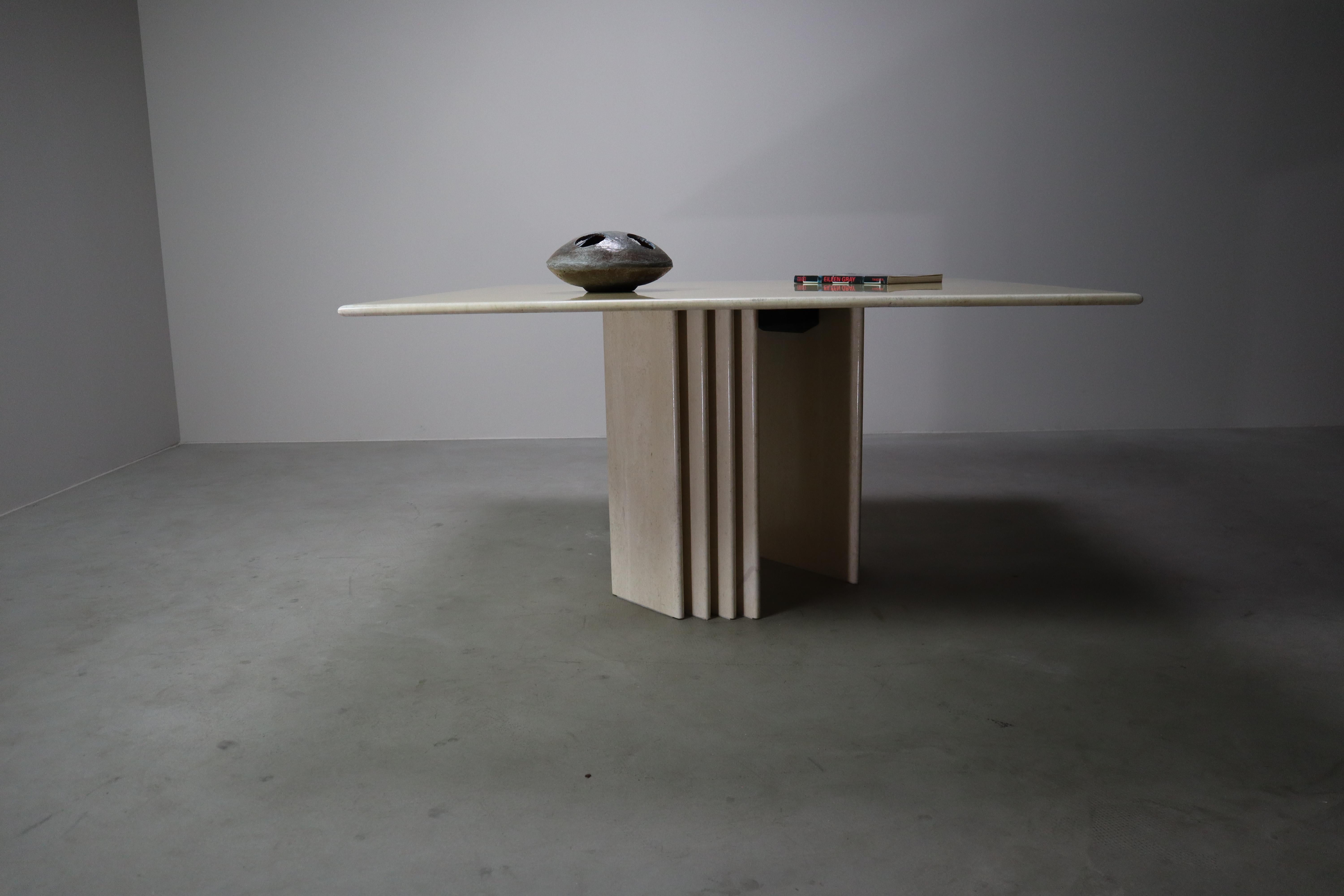 Esta llamativa mesa de comedor cuadrada de travertino es un refinado ejemplo del diseño escultórico italiano de finales de la década de 1960. La mesa presenta una base monumental compuesta de aletas verticales y planos planos, una audaz estructura