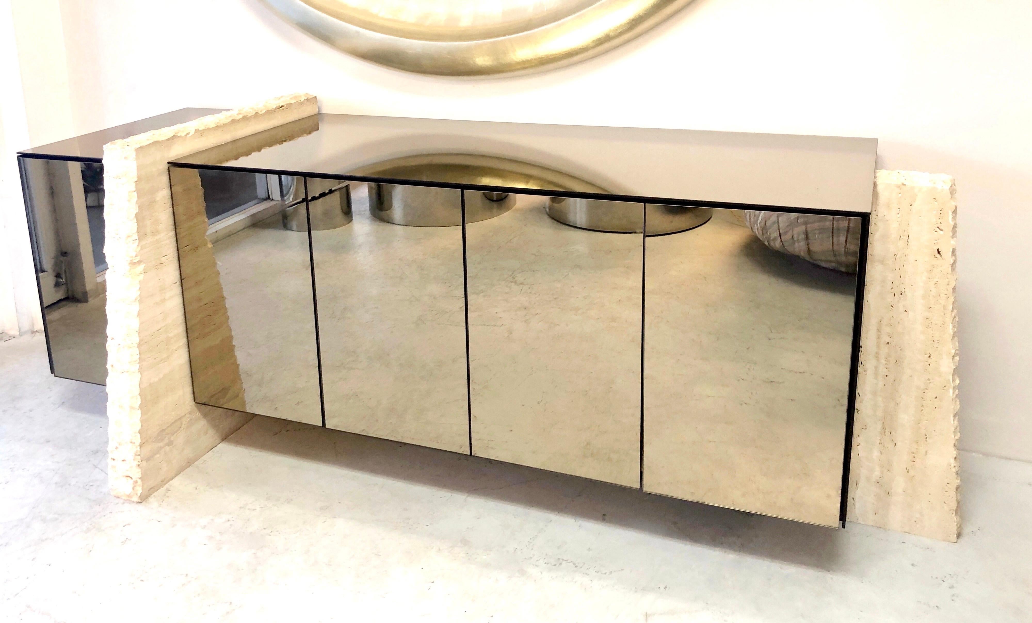 Spectacular console. The body is clad in bronze mirror, and supported on thick pieces of travertine marble. 5 doors conceal adjustable shelves.
The actual cabinet is 20.5” deep. The travertine slab is 27” at the floor.