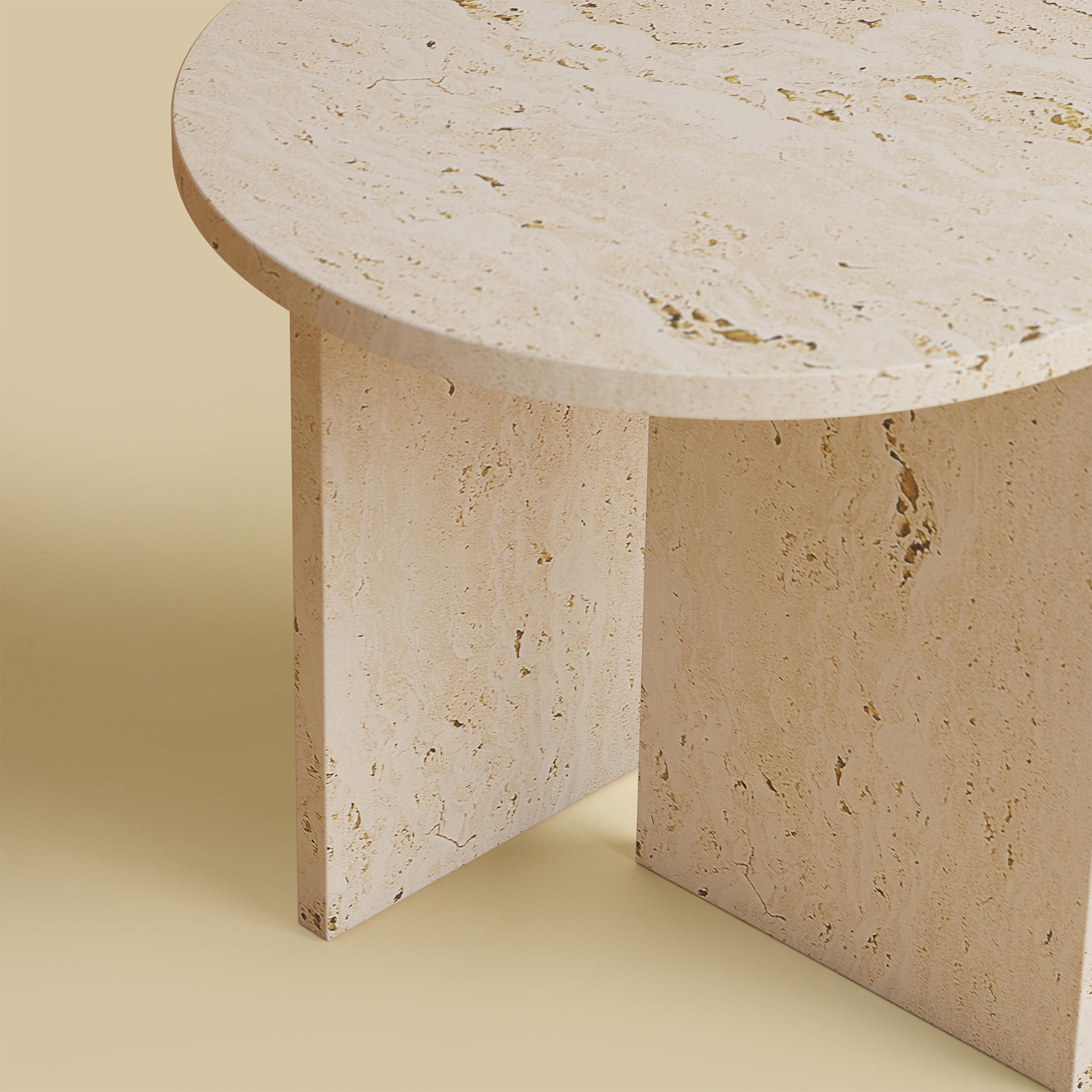 La table basse Kyushu est entièrement réalisée en travertin romain. Le plateau est circulaire et mesure 45 cm de diamètre. Les pieds sont constitués de deux plaques de marbre dont une partie est incrustée sur le plateau, ce qui constitue un détail