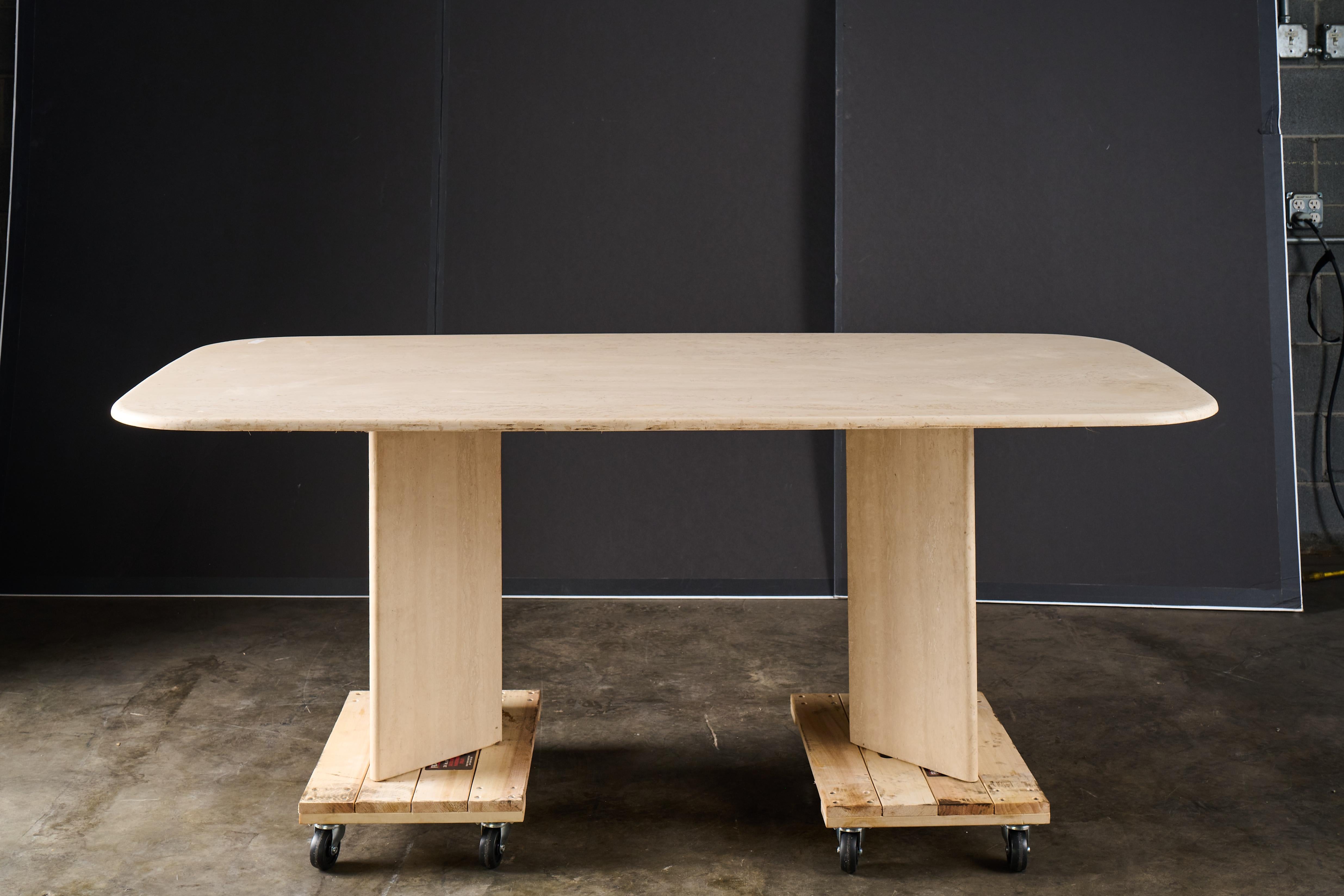 Table de salle à manger ou de conférence en travertin, Italie, vers les années 1970-80. Le plateau rectangulaire aux angles doucement arrondis repose sur deux socles sculpturaux en dalles. La tonalité neutre et chaude et les veines naturelles de la