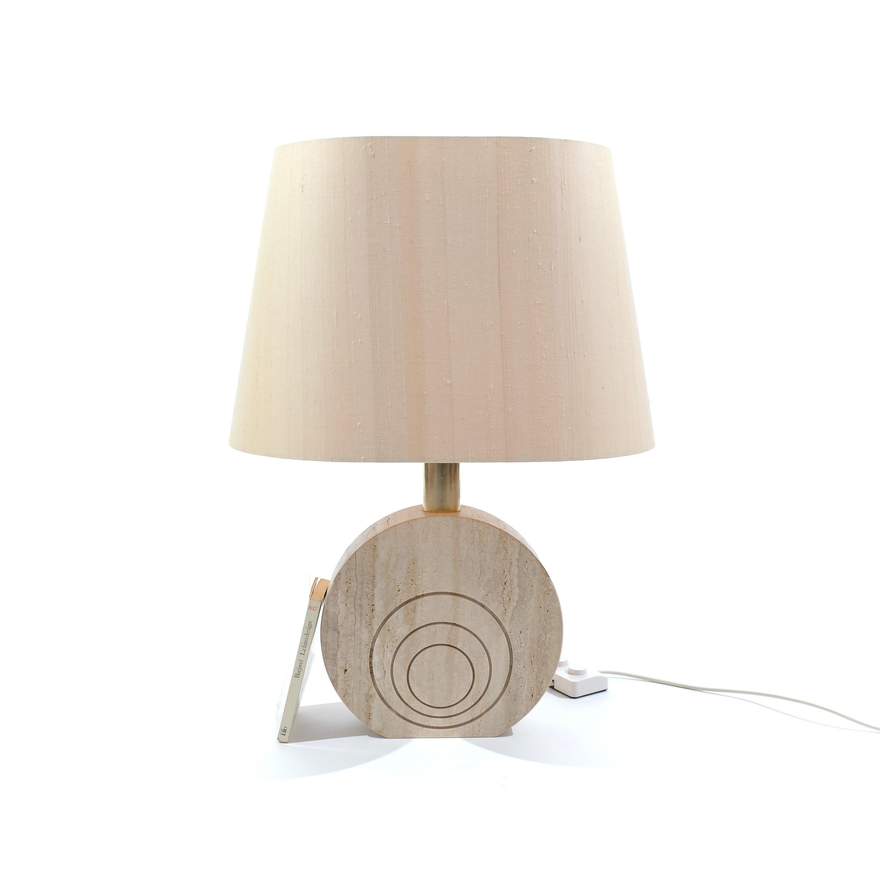 A thick disc of travertine, topped by a brass cylinder supporting a large cotton lampshade.

This shows all the refinement of Italian design. Noble materials used generously in perfect proportions to result in a delicate lamp with natural