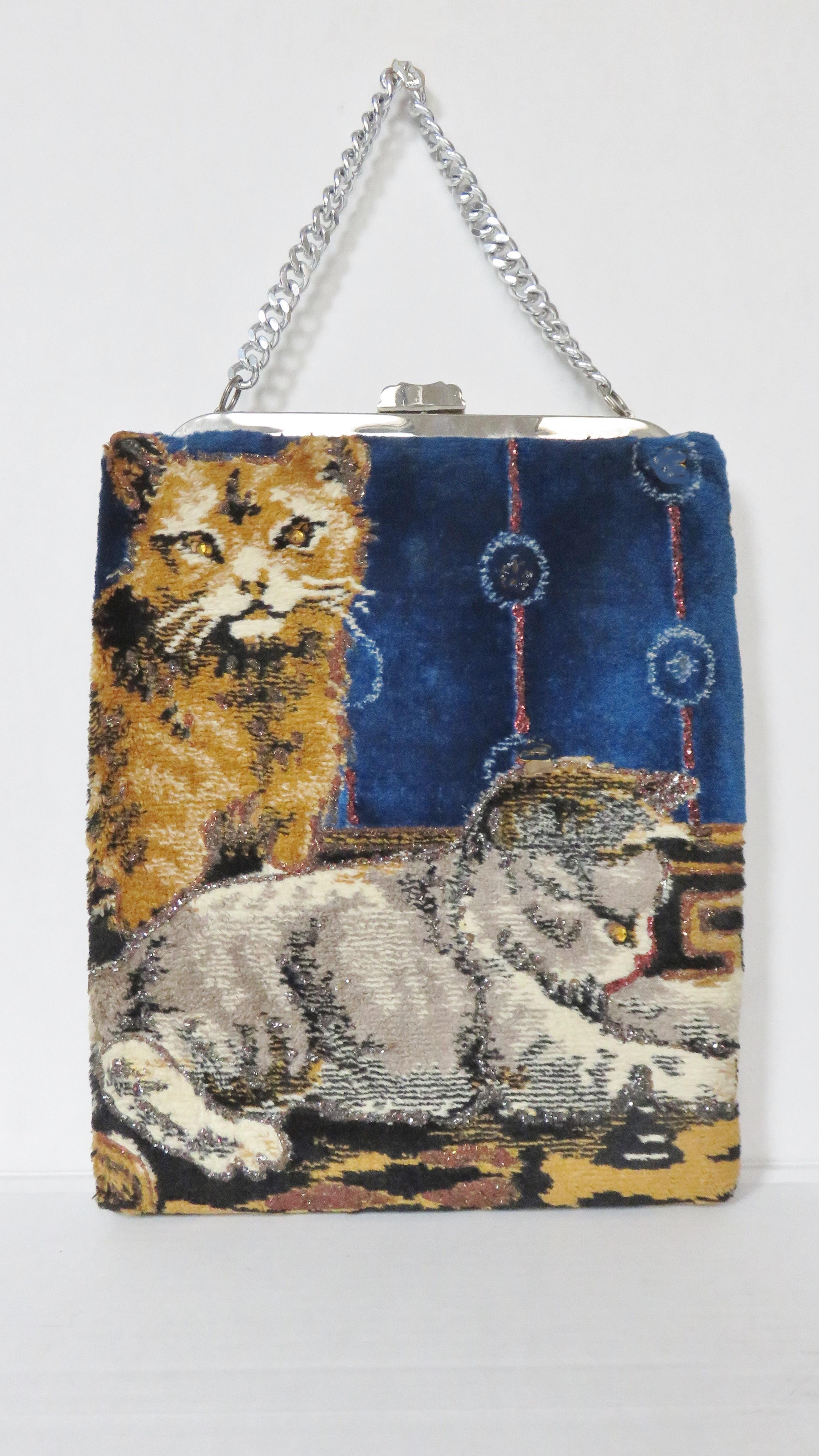 A great carpet bag from Tre-mor with cats on both sides.  It has a top silver chain handle, frame, clasp closure plus images of 2 cats on a blue background on each side. On one side the images are subtly outlined and have gold rhinestone eyes, the