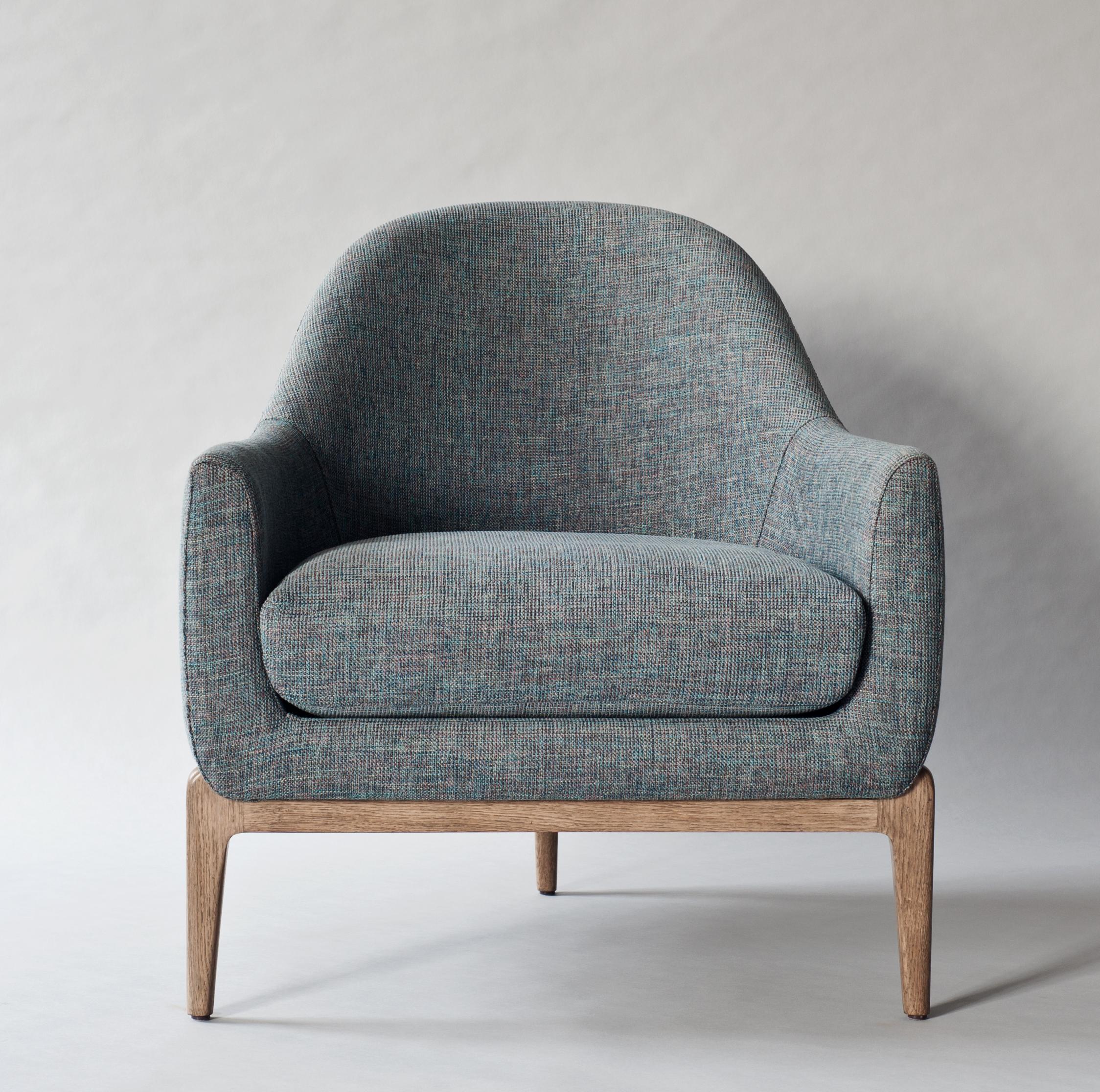 In the Treble Side Chair, a gently curved back gives way to streamlined armrests, embracing the body and framing a loose seat cushion. The fully upholstered form rests atop a tripod base of hand-crafted solid oak, with tapered legs that mirror the