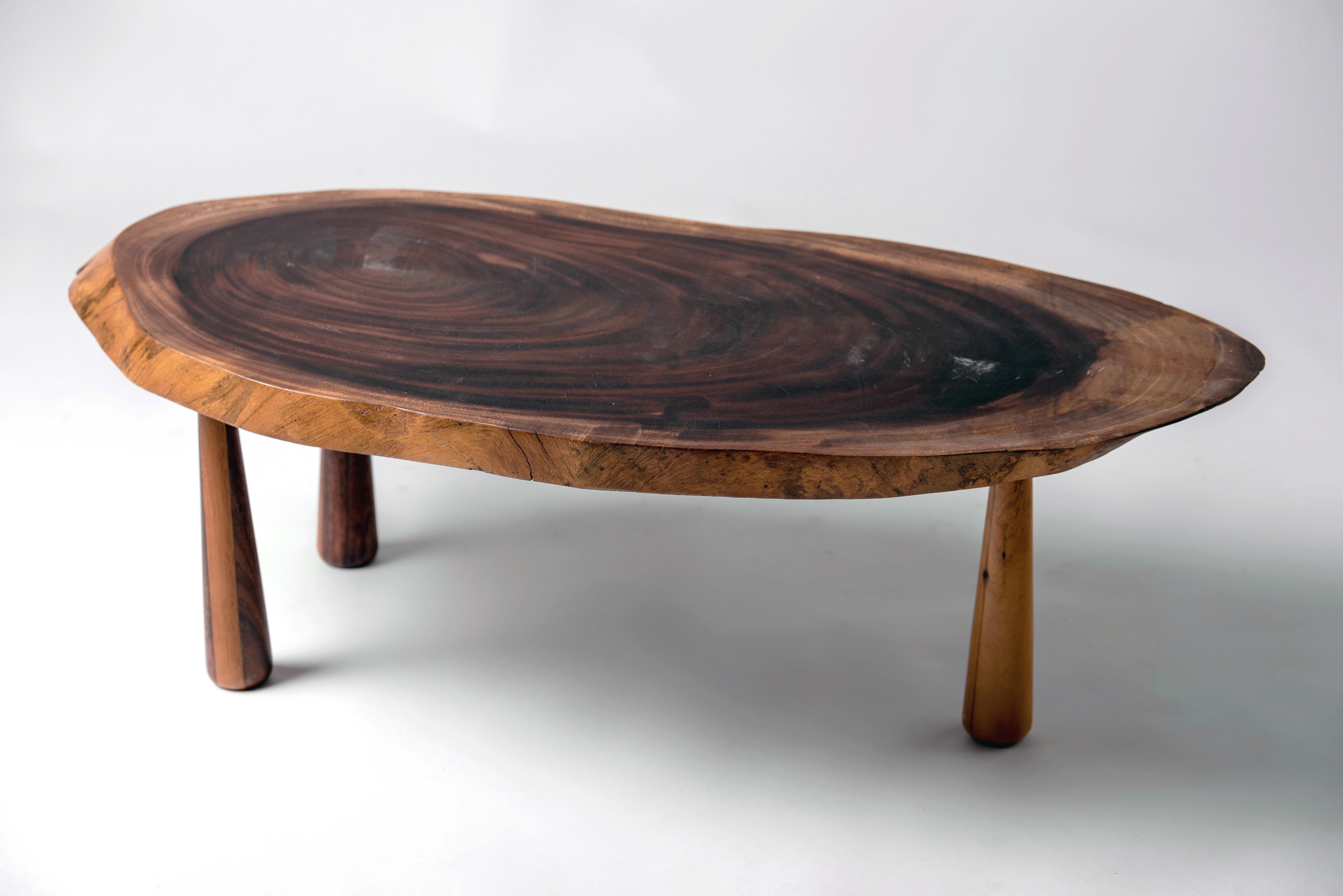Table basse intéressante. Un plateau fait d'une tranche d'arbre repose sur trois pieds coniques faits de deux bois, clair et foncé.