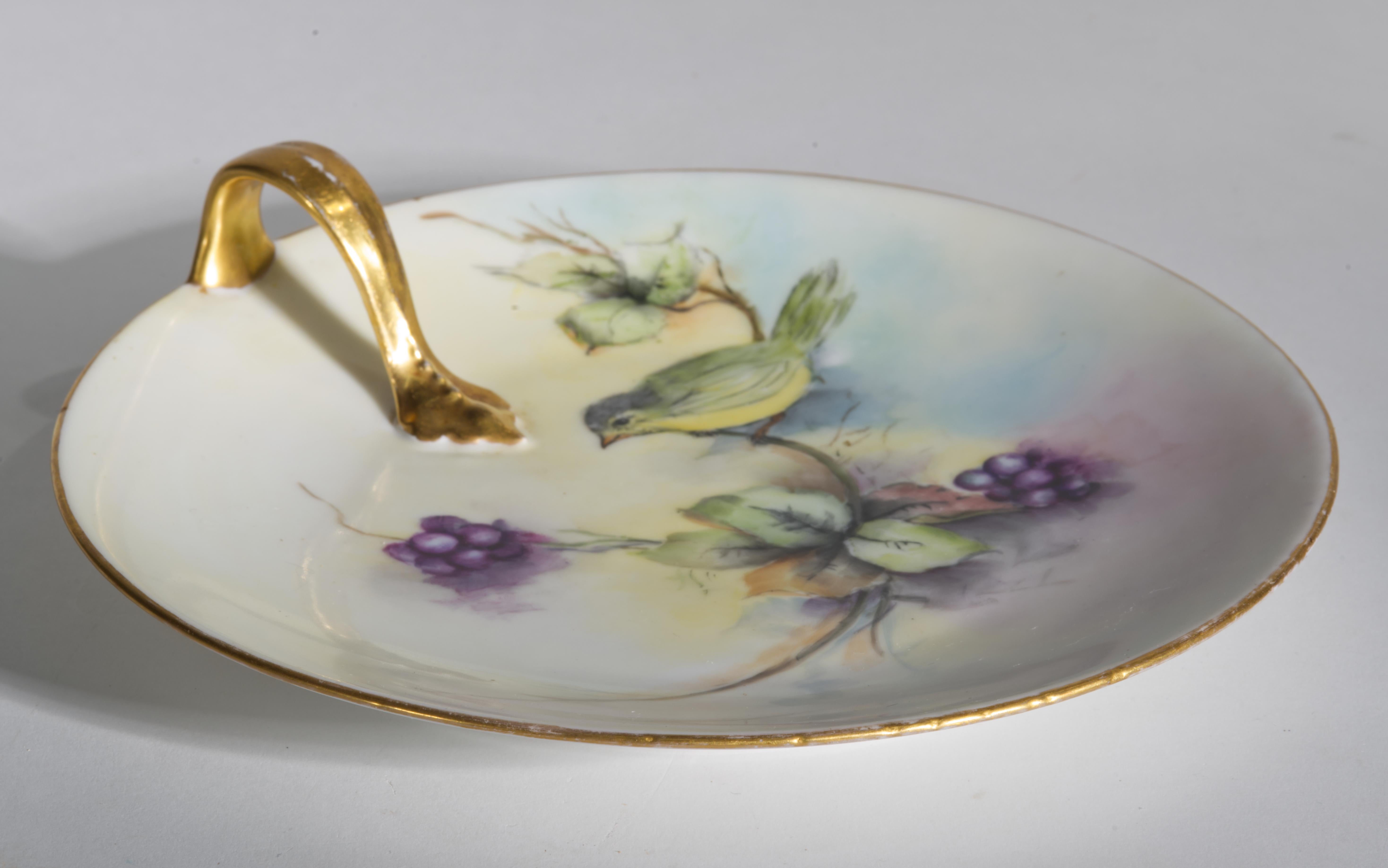 Plateau ancien en porcelaine avec poignée arrondie présentant un décor peint à la main d'un petit oiseau, assis sur une branche avec des feuilles et des baies violettes. L'arrière-plan est réalisé dans des couleurs pastel douces, suggérant l'air