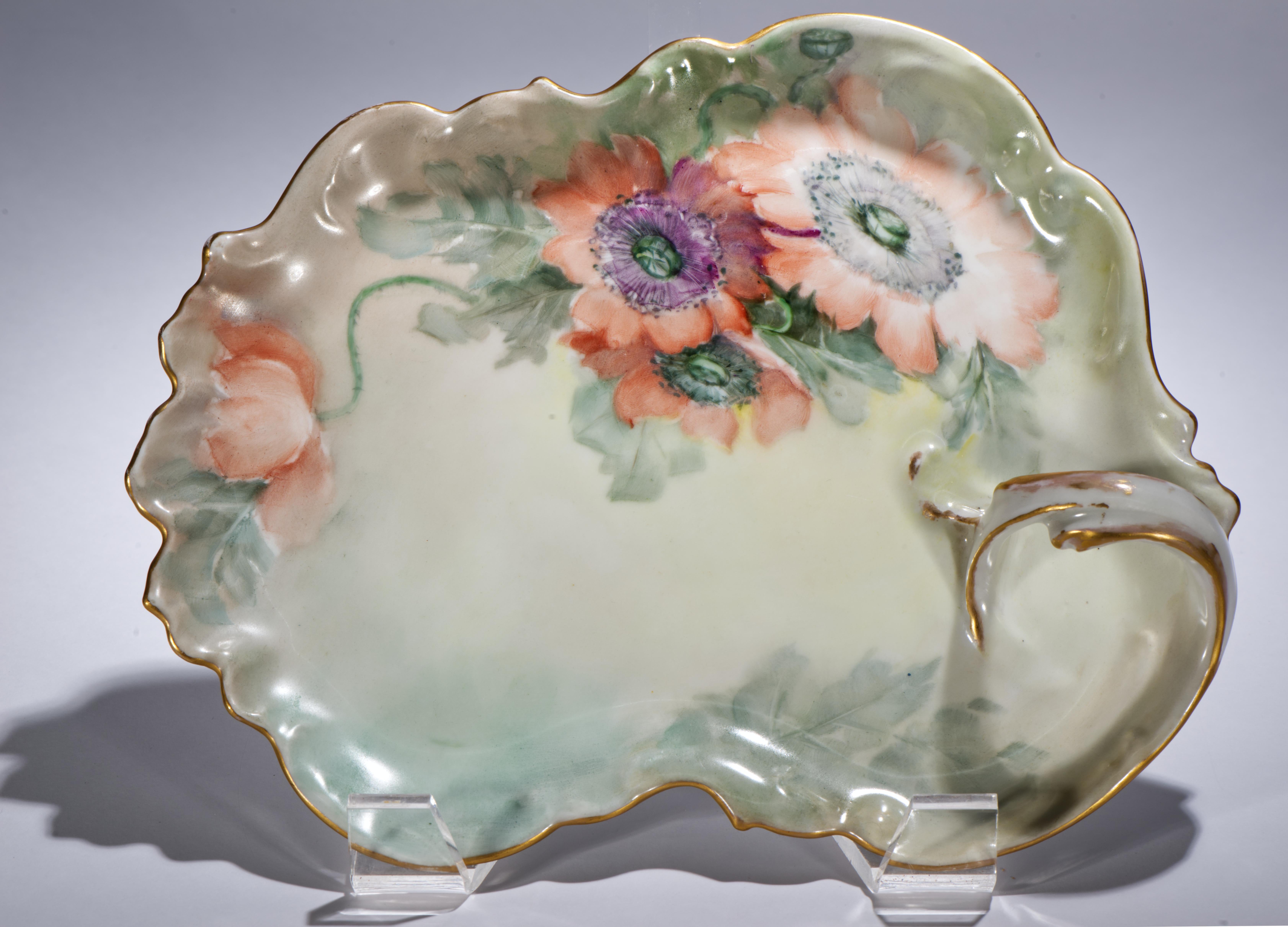L'antico vassoio in porcellana con manico arrotondato presenta un decoro dipinto a mano con fiori di papavero e foglie. Lo sfondo è realizzato con colori pastello tenui, che suggeriscono l'aria aperta e altri rami dietro il motivo principale. Il