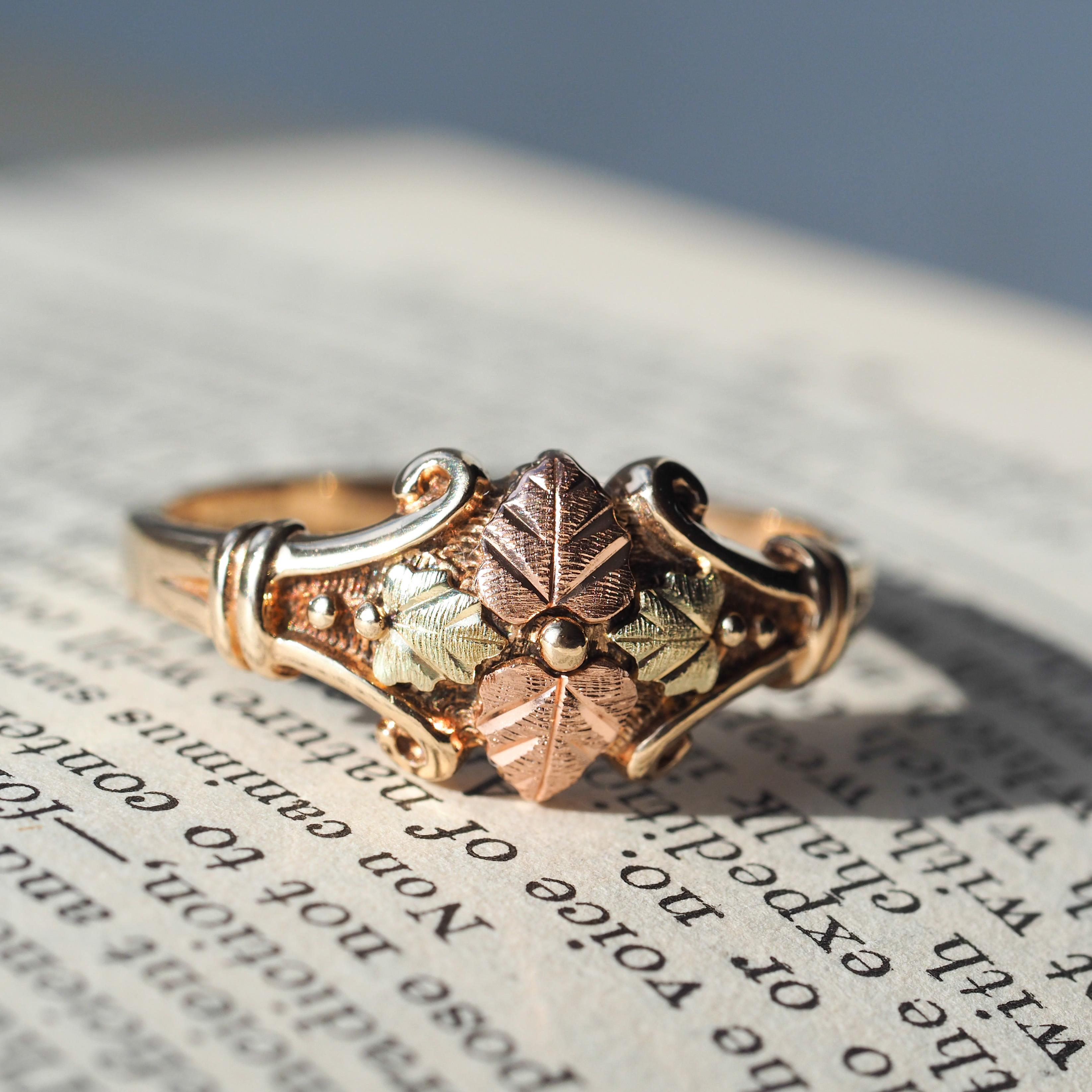 We are delighted to offer this beautiful tri-colour 9ct gold vintage ring.

The ring features a understated yet intricate foliate design with fine engravings on the top leaves encompassed by a scrolled border which joins the shoulders of the