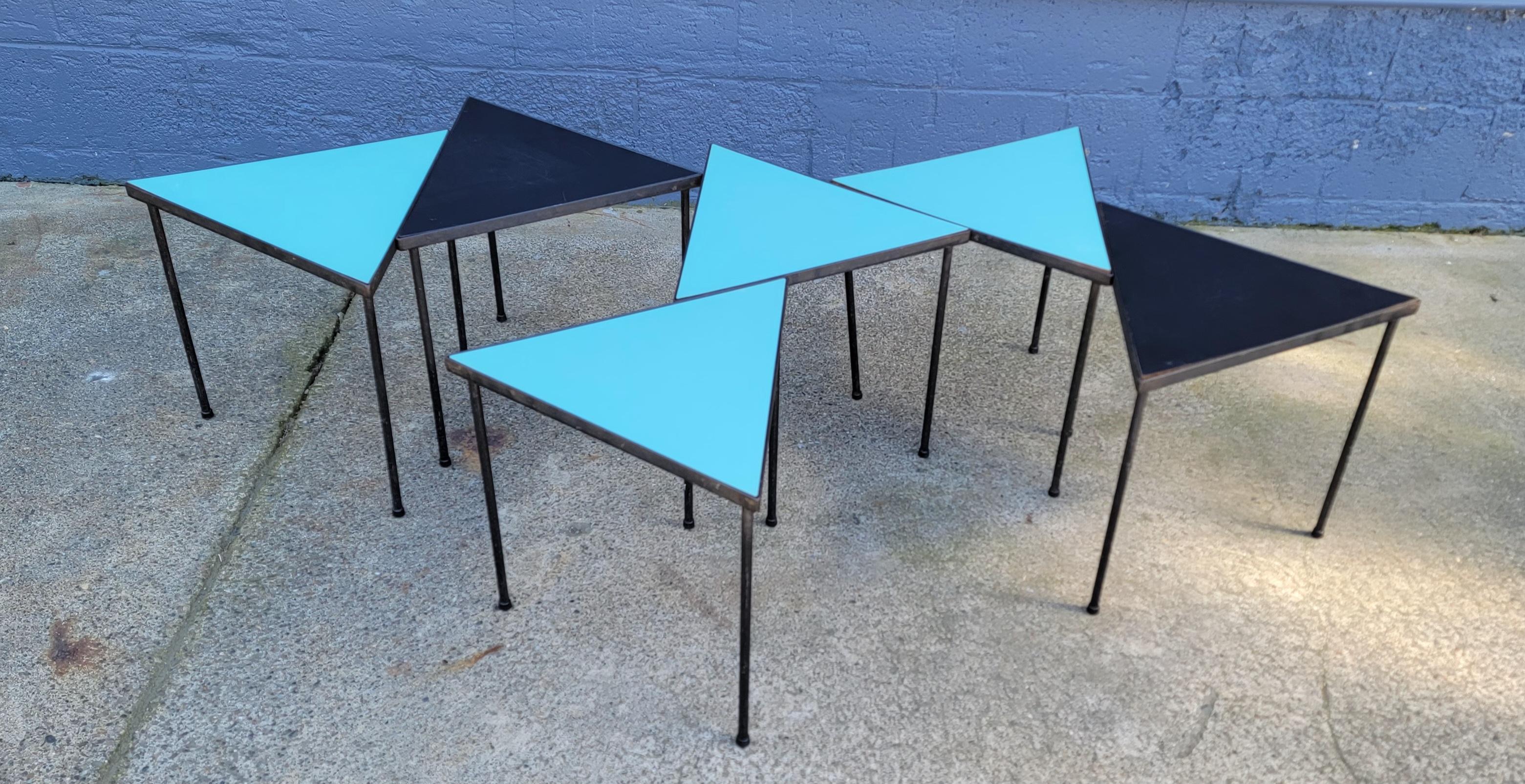 Eine Reihe von mehrfarbigen Tischen mit Eisenfuß und Formica-Platte, die ineinander verschachtelt oder modular aufgebaut werden können. Vier türkisfarbene Oberteile und zwei schwarze Oberteile. Diese Tische können ineinander gestapelt oder in