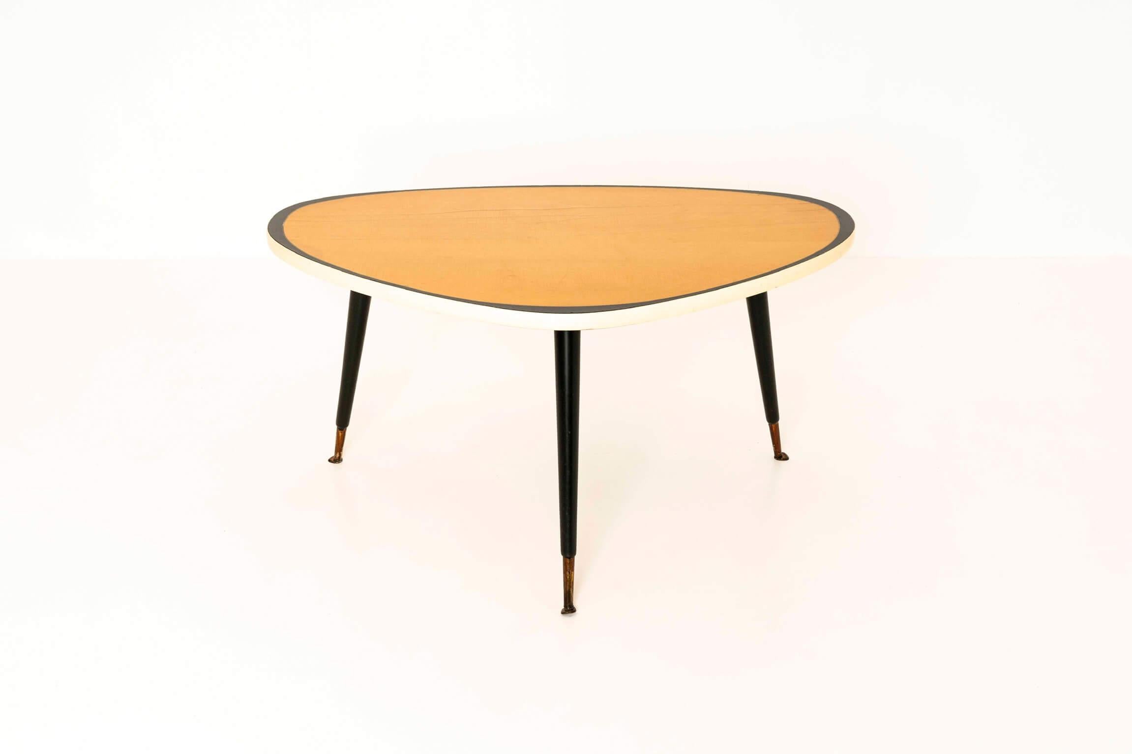 Nice triangular vintage coffee table from Germany, the 1950s. The table is manufactured in the style of, and could very well be by, Ilse möbel. The three black legs have brass feet. The table has a yellow top with, due to its age, stripes in the