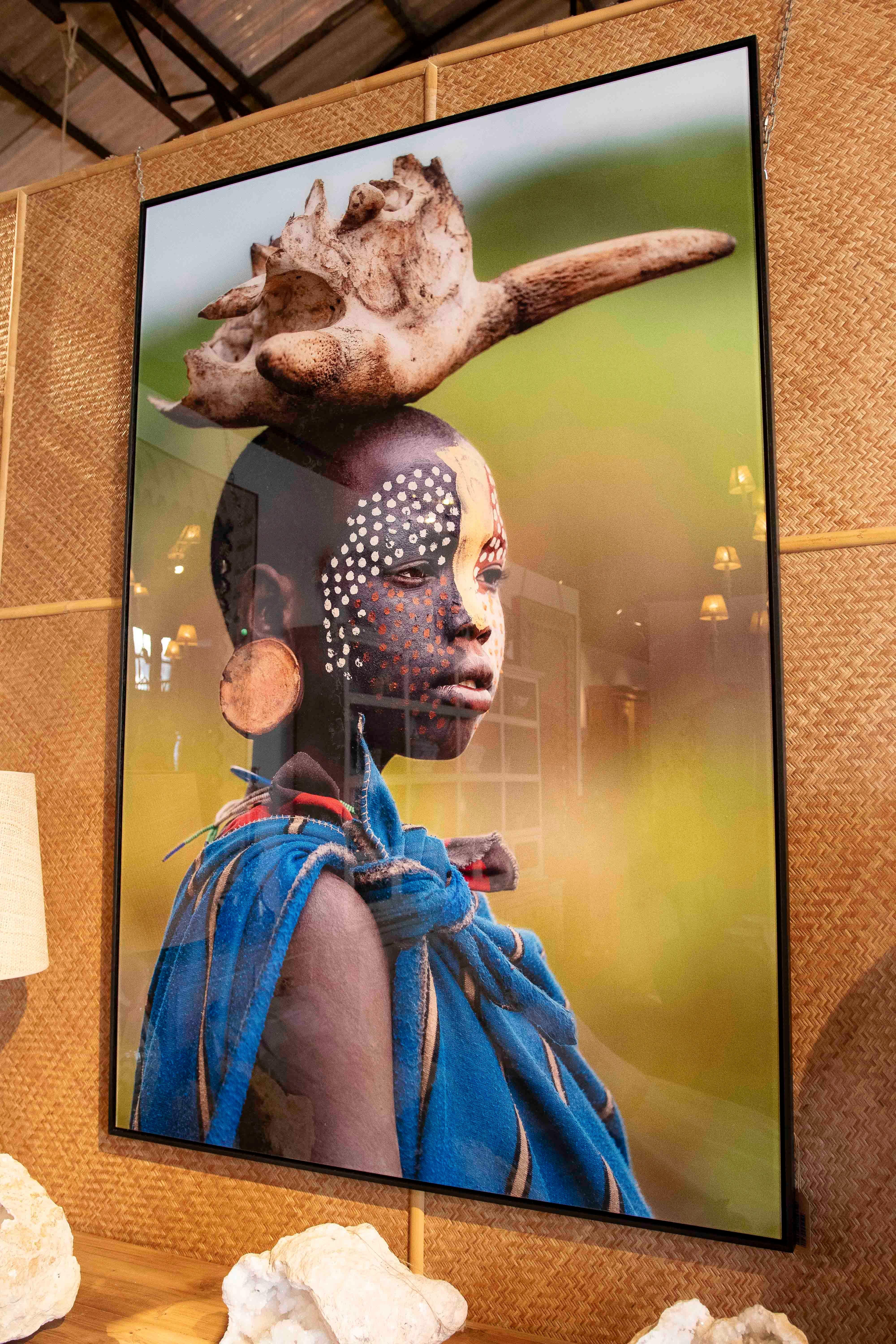 Striking photograph capturing the cultural essence of a young woman in an African landscape, wearing an elaborate headdress of natural bone and symbolic facial paintings in vibrant tones. The contrast between her skin, the details of her blue
