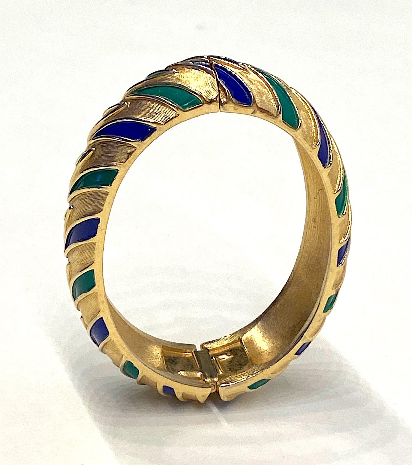 A lovely 1960s Trifari satin gold bangle with navy blue and green enamel stripe accents. The bangle is an oval shape with a light dome or curve measuring .25 of an inch thick. It has a hinge on one side and a safety clasp on the opposite side. The