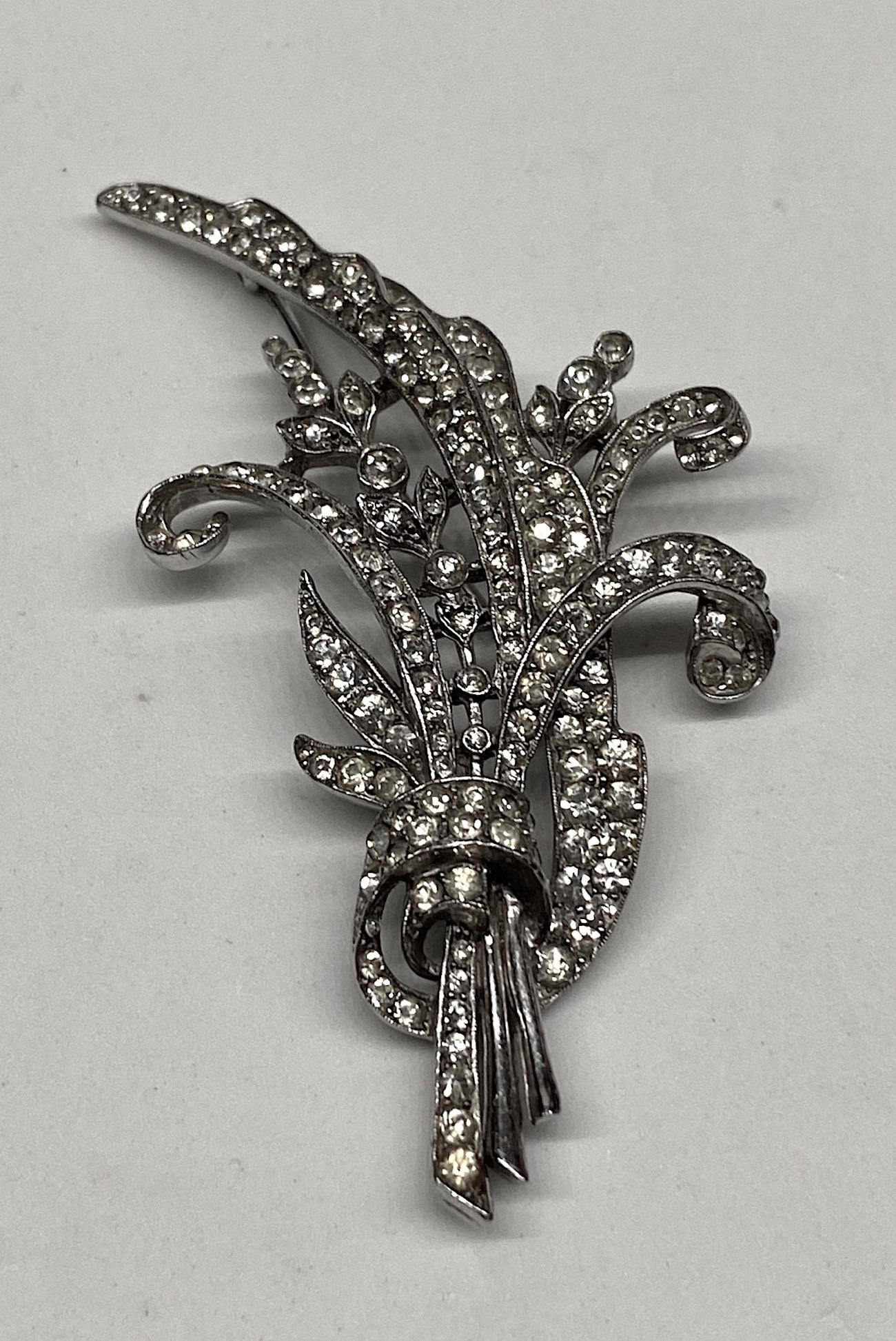 A large rhodium plate and rhinestone floral spray in the Art Deco style by Trifari. The design is a bouquet of long spirals, a long leaf and branch of tiny two petal flowers all sweeping upwards. The brooch is 2 inches wide, 3.38 inches high and .5