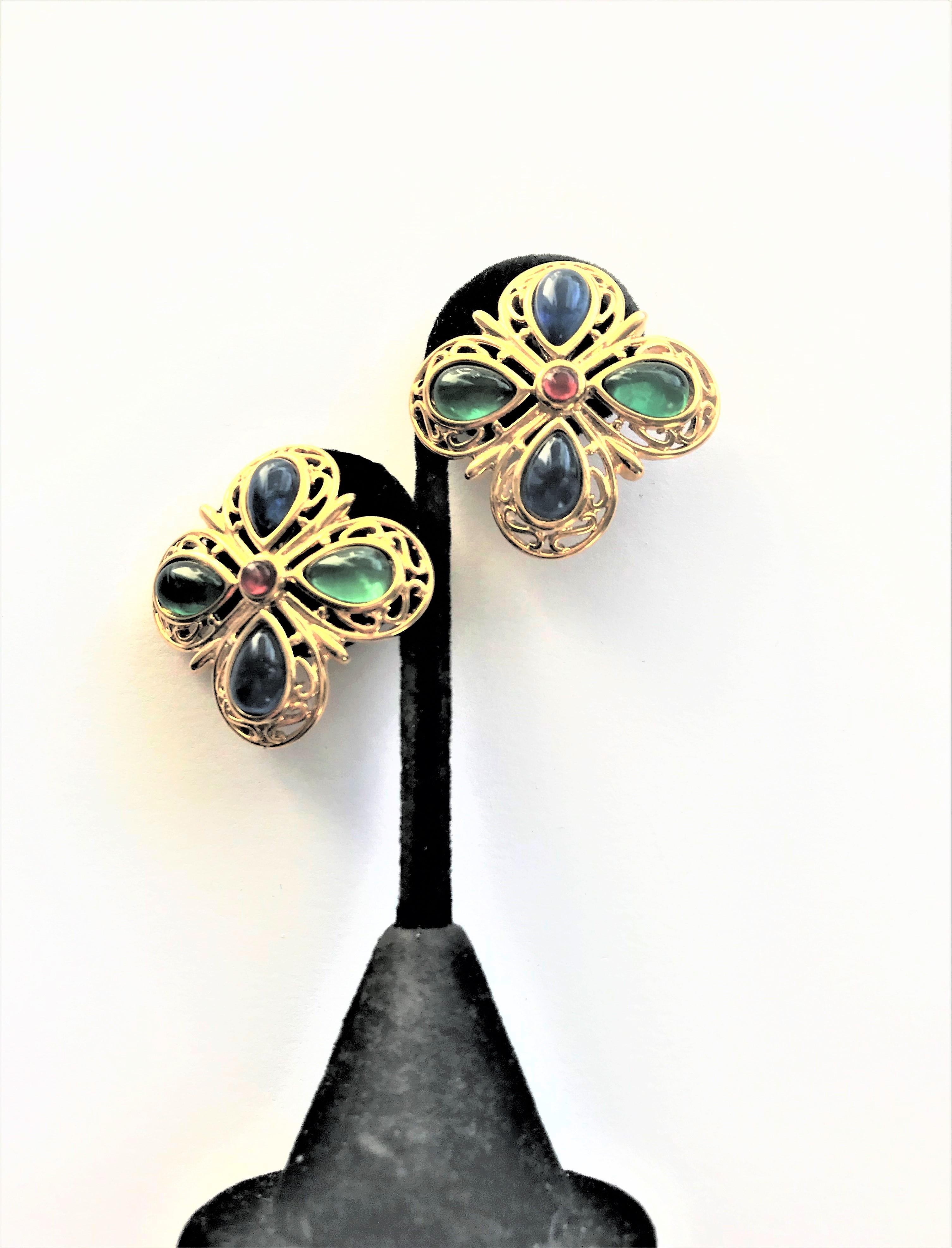 Trifari Ohrclips in Form von 2 4-blättrigen Kleeblättern mit blauen und grünen  Glastropfen. Eingefasst mit durchbrochener Goldarbeit. In der Mitte ein kleiner roter Glasstein. Auf der Rückseite der Clips Klammer Trifari Unterschrift. 
Messung:  3,5