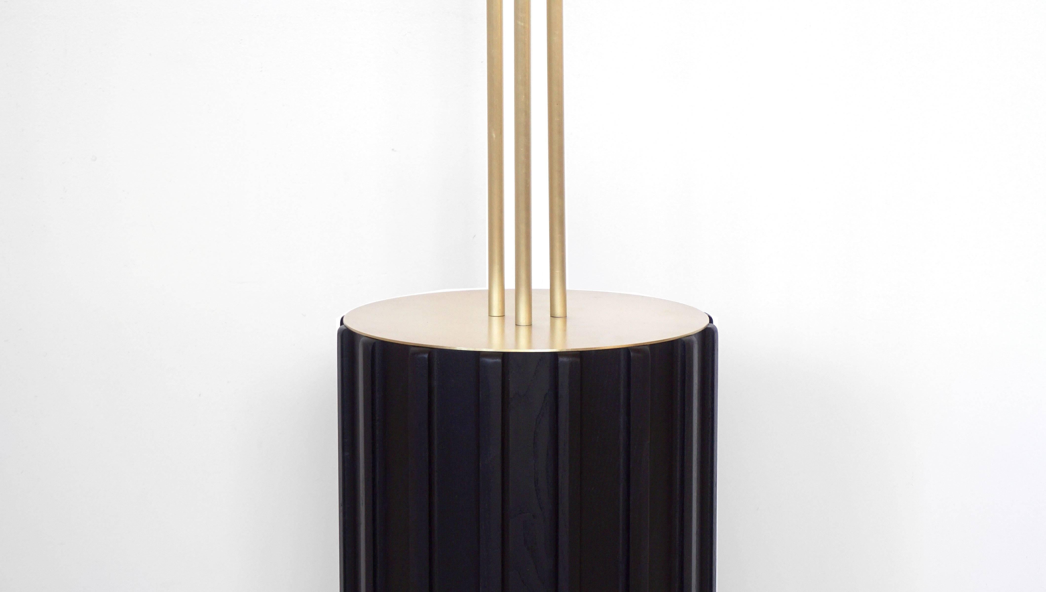 Trillium floor lamp is made of brass and blackened ashwood. Three rings sit at eye level, each with a light source hidden within, between two brass discs. The light seeps out from the discs and is bounced off the interior of the rings, with a soft