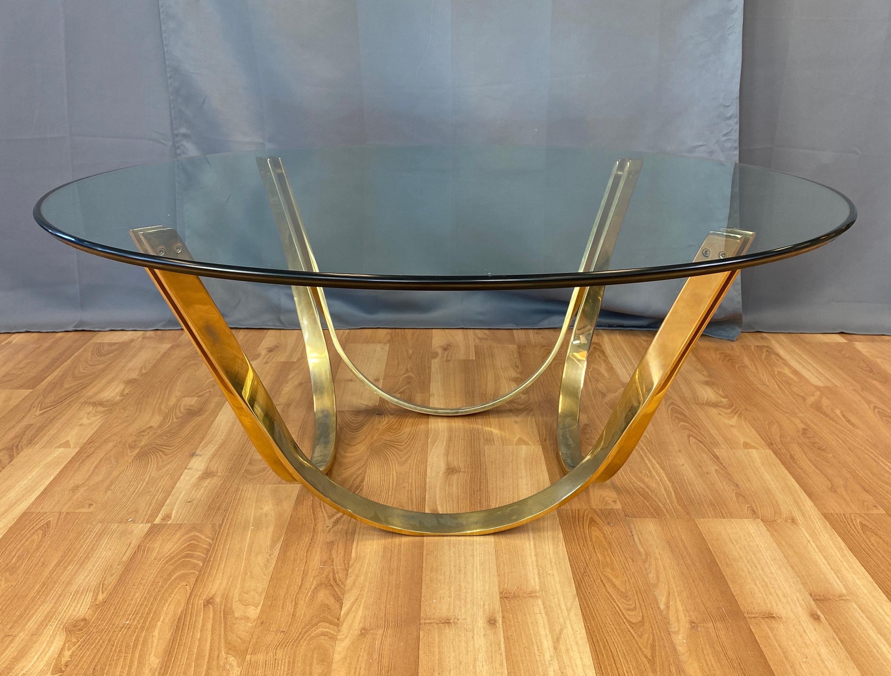 A 1970s brass-plated steel and glass top round coffee table by TriMark, done in the style of Roger Lee Sprunger for Dunbar Furniture.

Handsomely executed base comprised of four brass finish flat bar steel ribbons that elegantly curve, twist, and